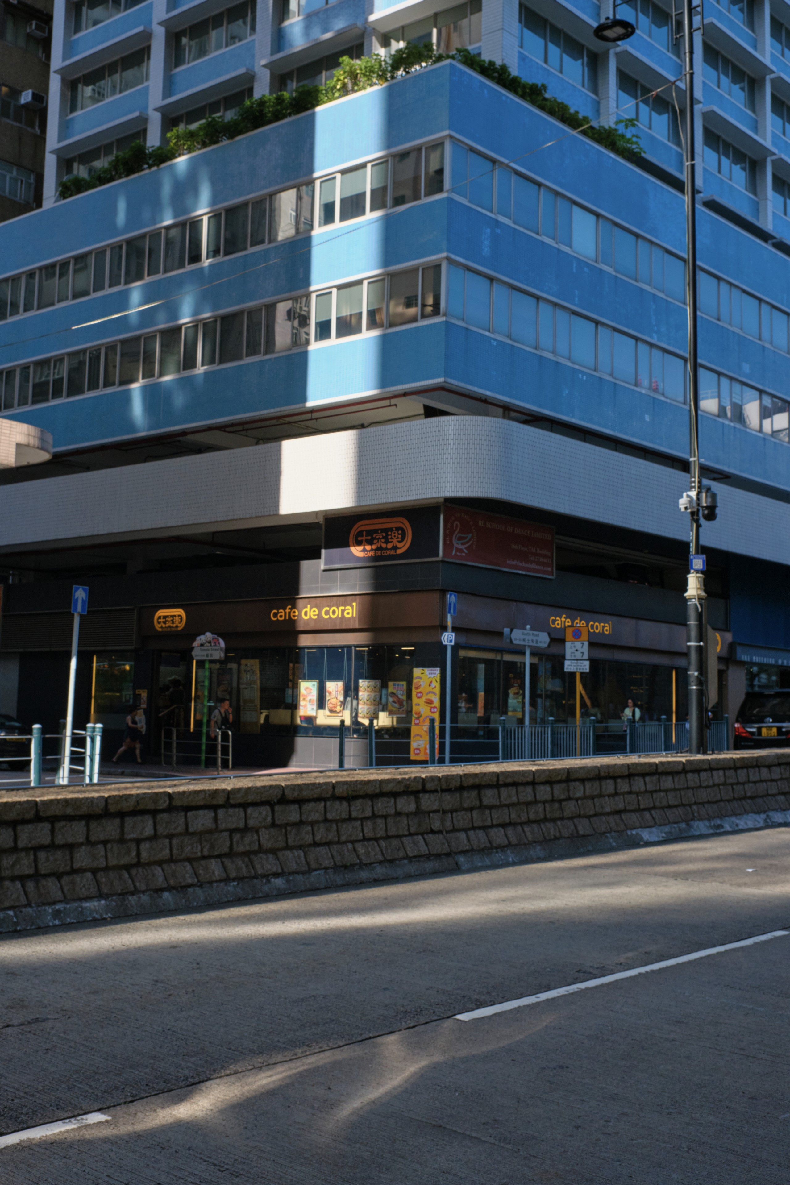 Corner of a blue building with a ground-floor shop featuring orange signage. Street in foreground with sunlight and shadows