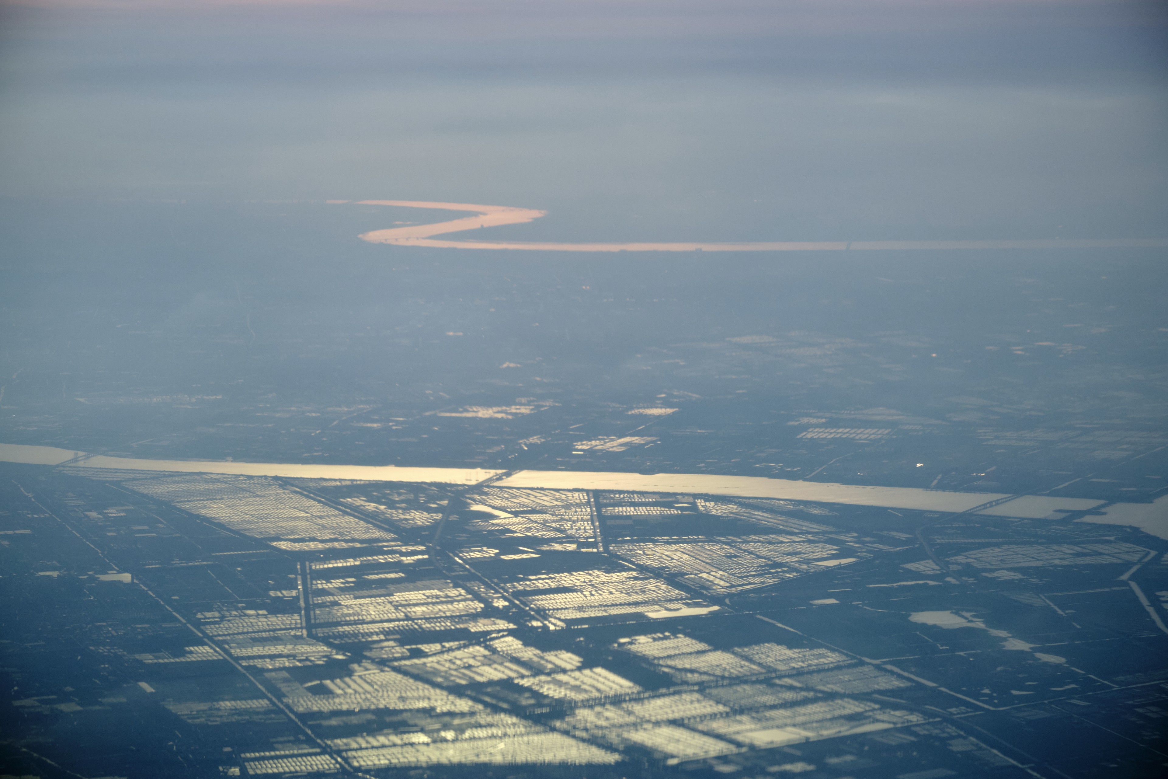 Aerial view of a reddish, winding river flowing through a hazy, snow-dusted landscape featuring rectangular fields and a wider river
