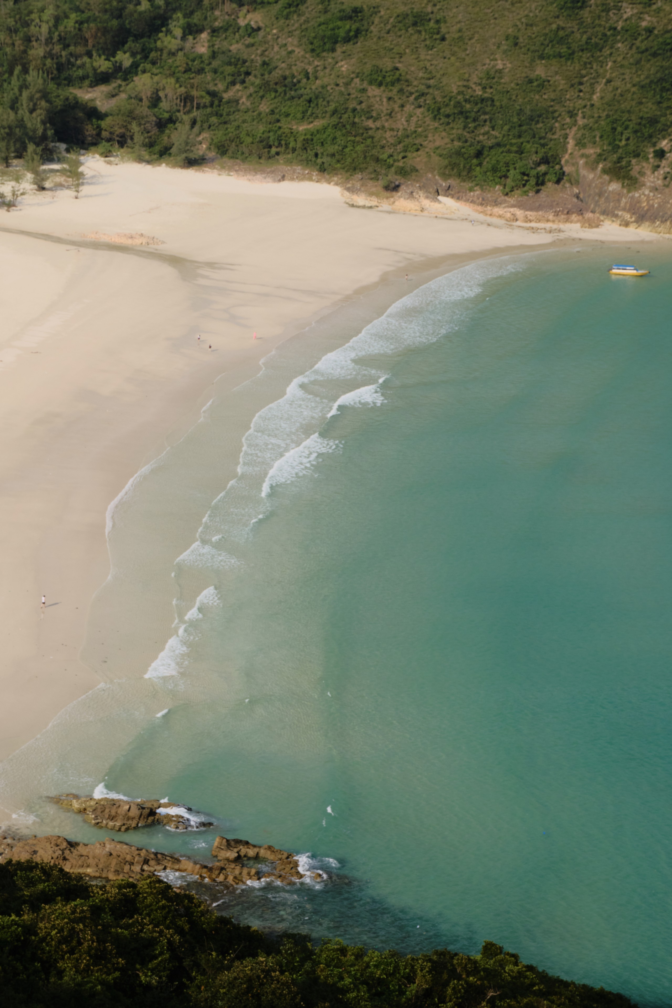 Aerial view of a crescent white sand beach meeting turquoise ocean water with gentle waves, framed by green hills and rocky coastline