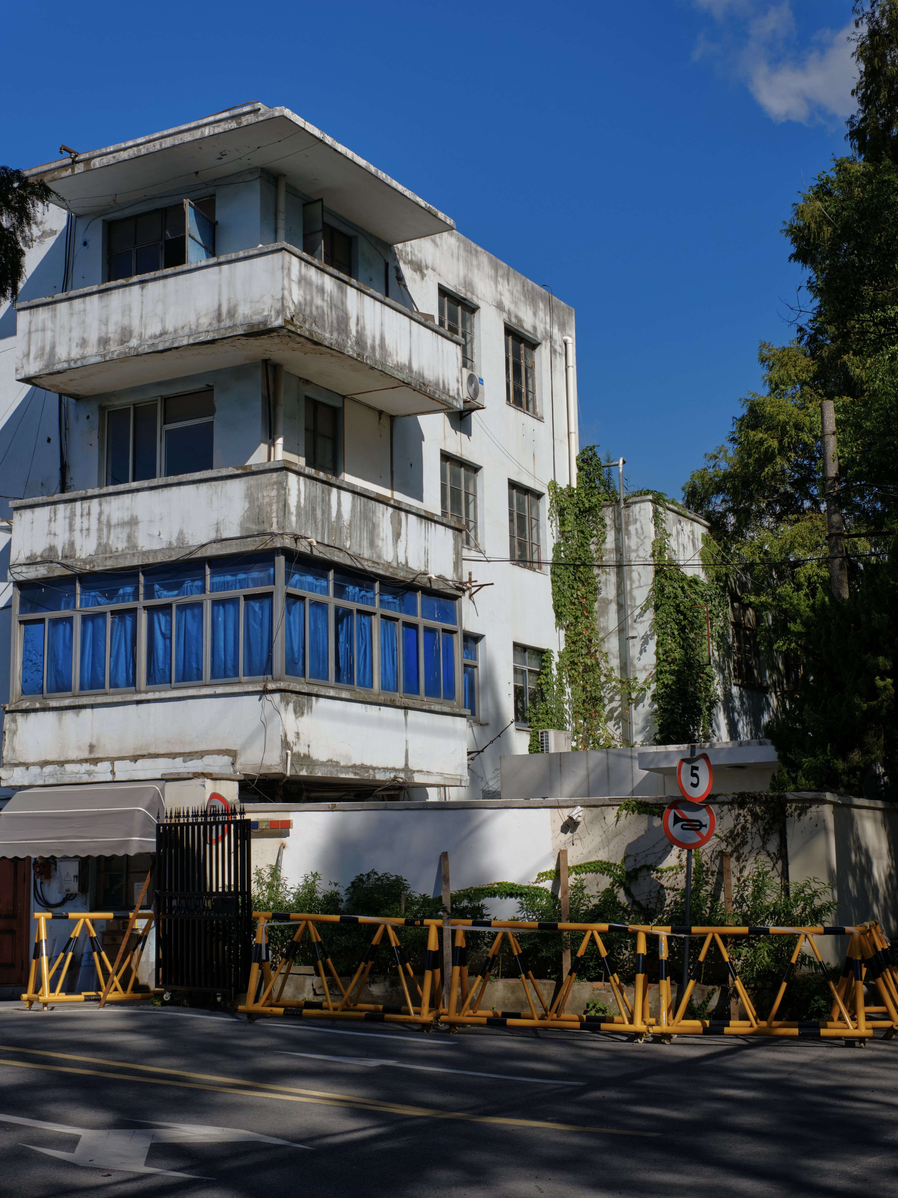 White three-story building with multiple balconies and prominent blue windows on the second floor, bordered by a white wall and numerous yellow barriers on a dark street, under a clear blue sky