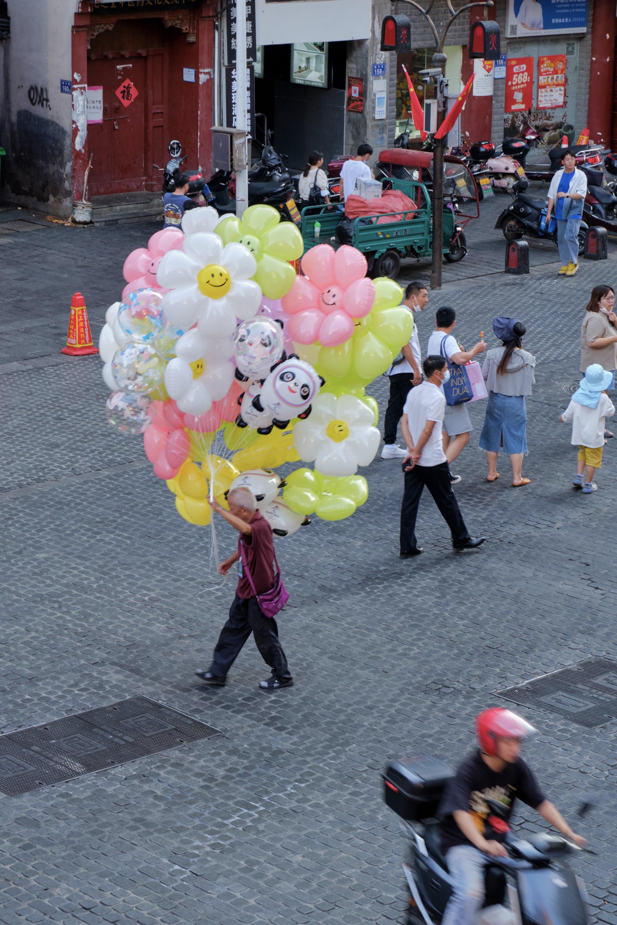 A person walks across a paved street carrying a large cluster of flower and panda-shaped balloons. Other pedestrians, parked scooters, and buildings are visible in the background