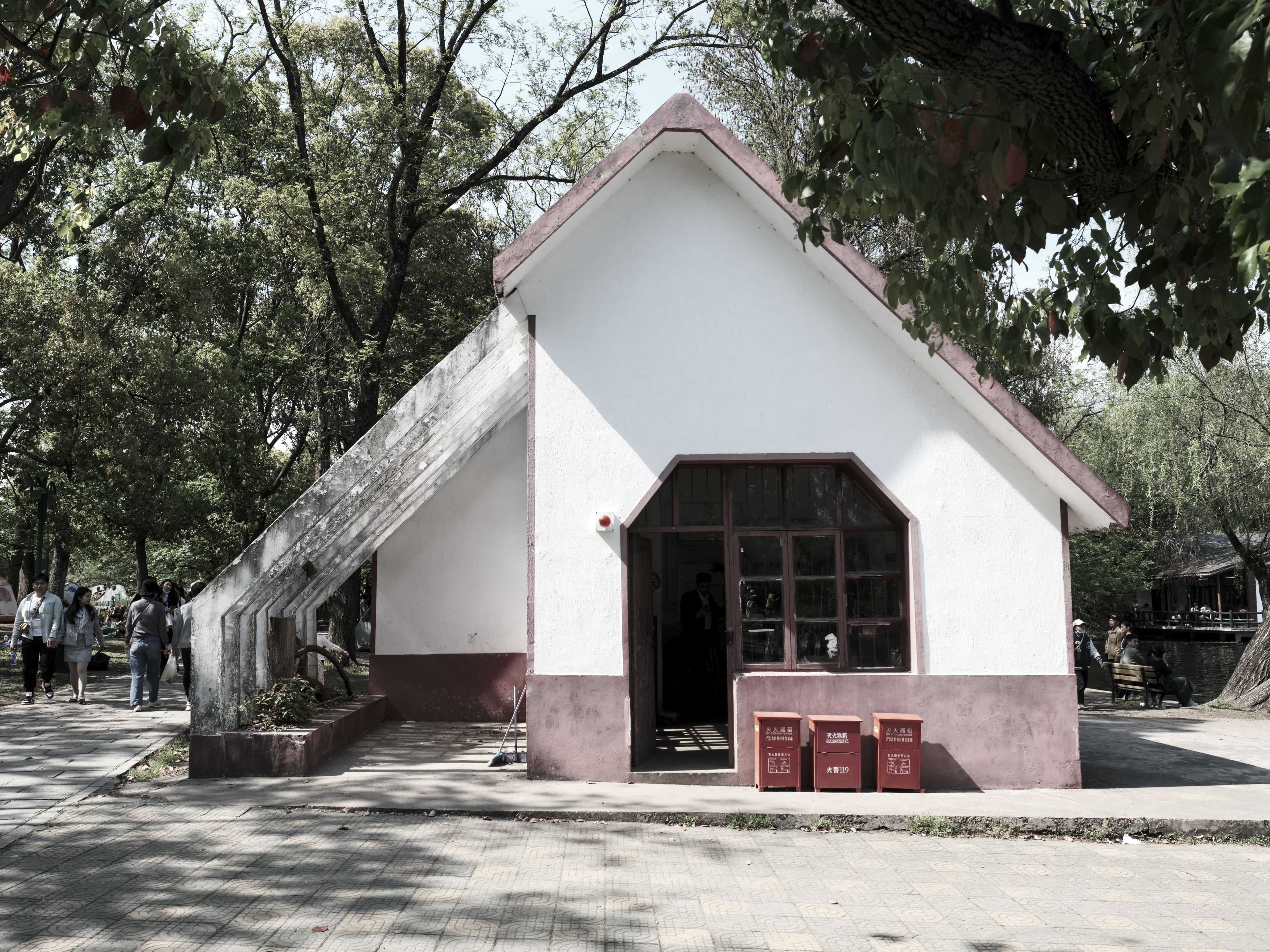 Small white building, red base, pitched roof, attached awning, two red containers, surrounding trees