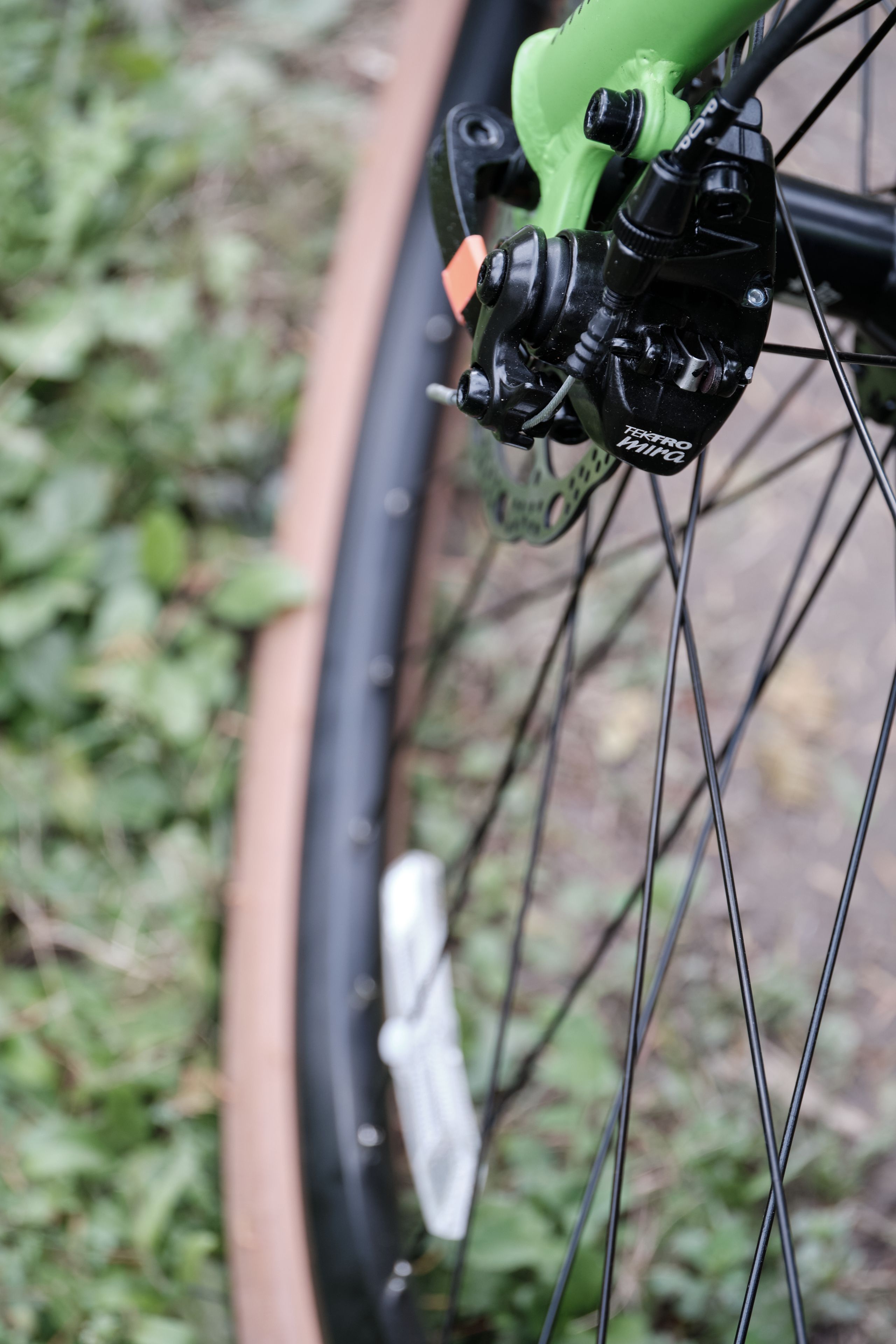 Close-up bicycle front wheel, disc brake, green fork, tan tire, spokes, blurry grass