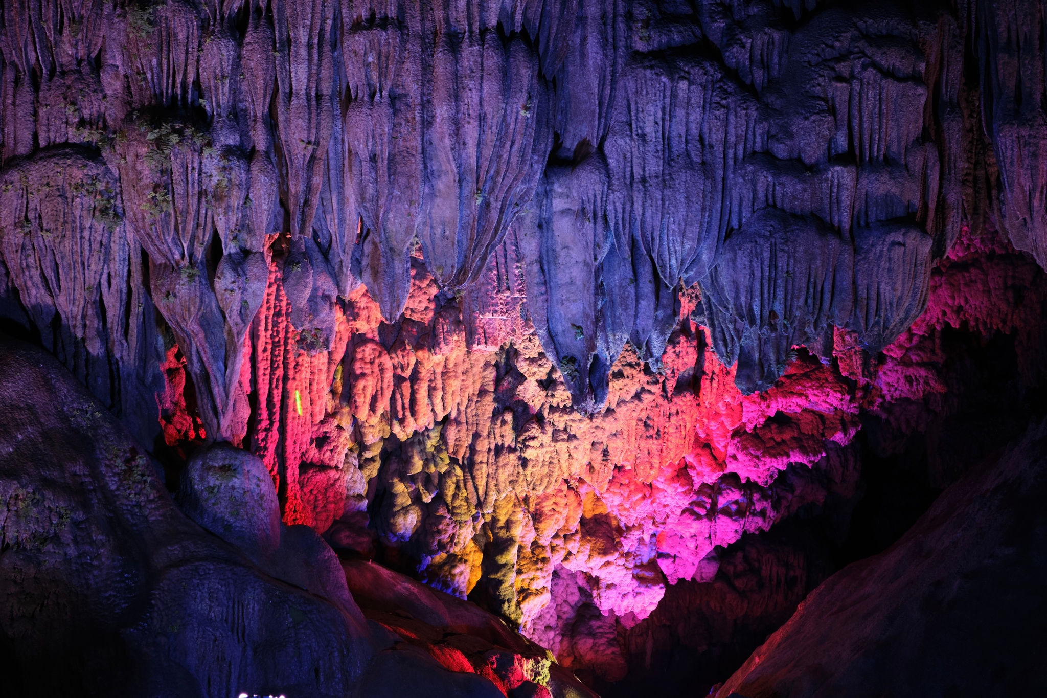 Cave interior with colorful stalactites and stalagmites illuminated in purple, blue, red, pink, and yellow light