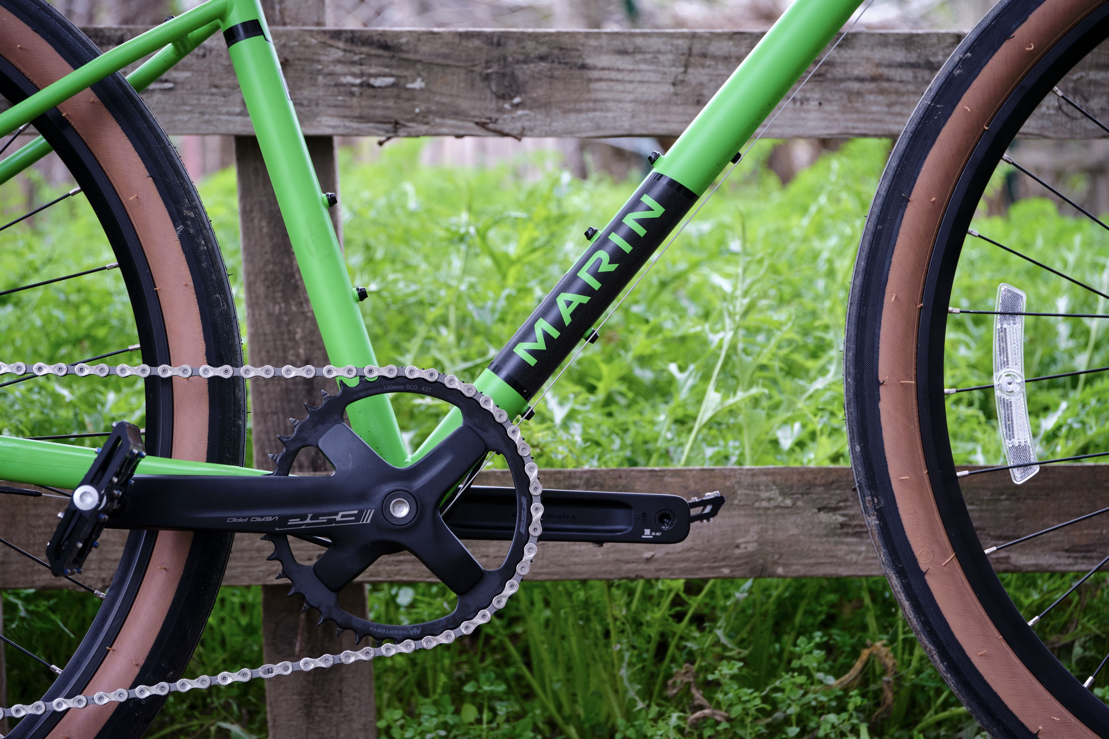 Close-up of a bright green bicycle frame, crankset, chain, pedals, and parts of wheels with brown sidewalls against a blurred background of green grass and a wooden fence