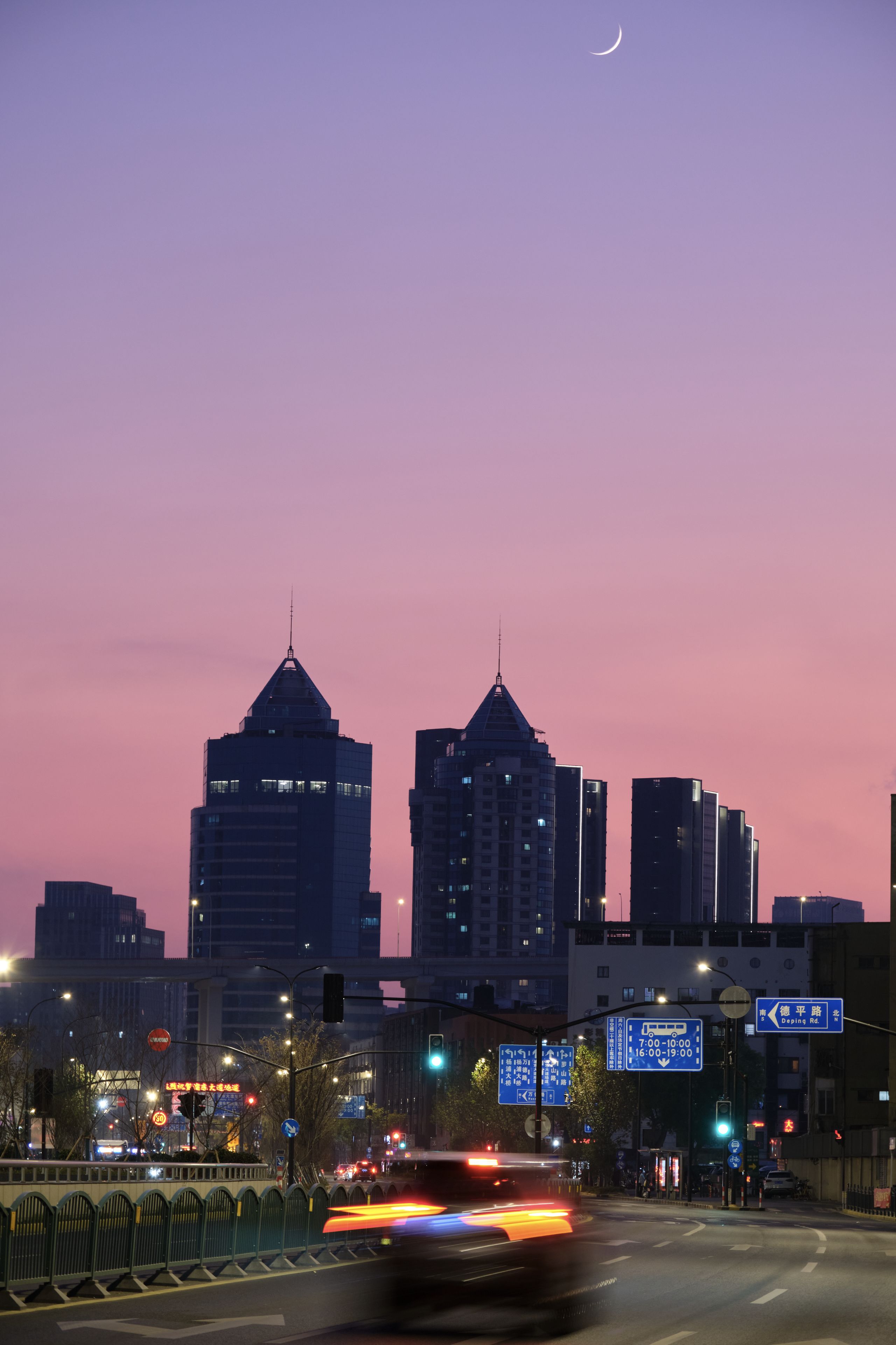 City skyline with tall buildings against a purple twilight sky, a crescent moon visible, and blurred car lights on a street below