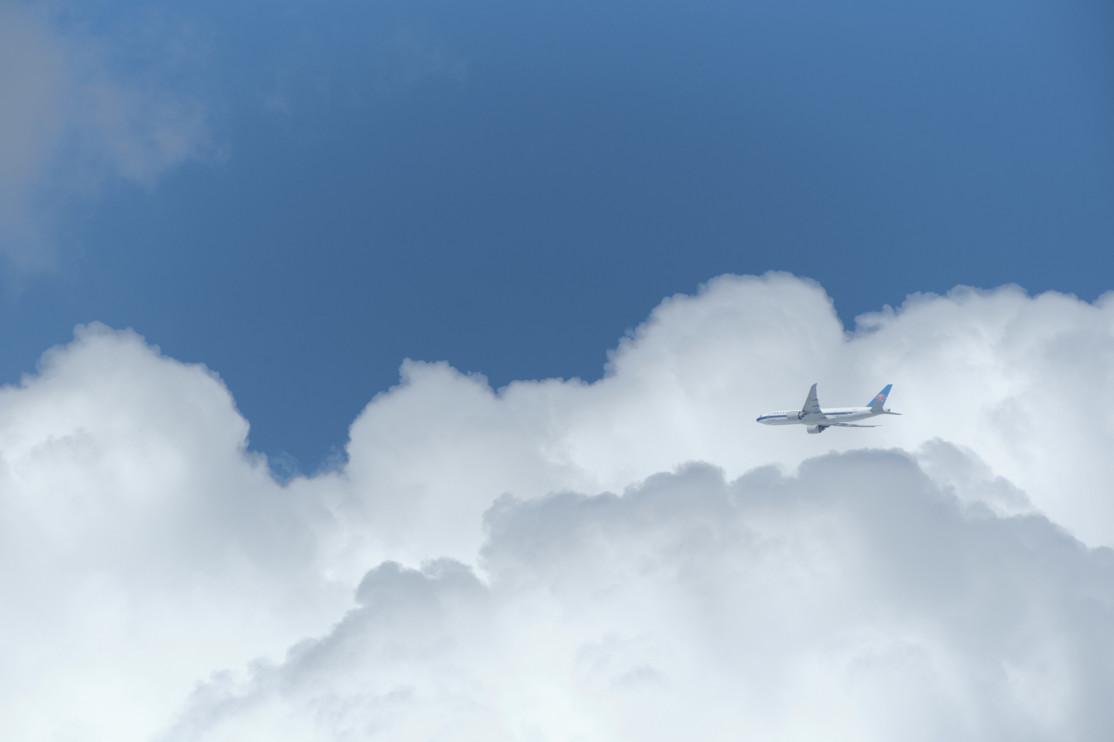 A white passenger airplane flies horizontally above a layer of fluffy white clouds against a clear blue sky