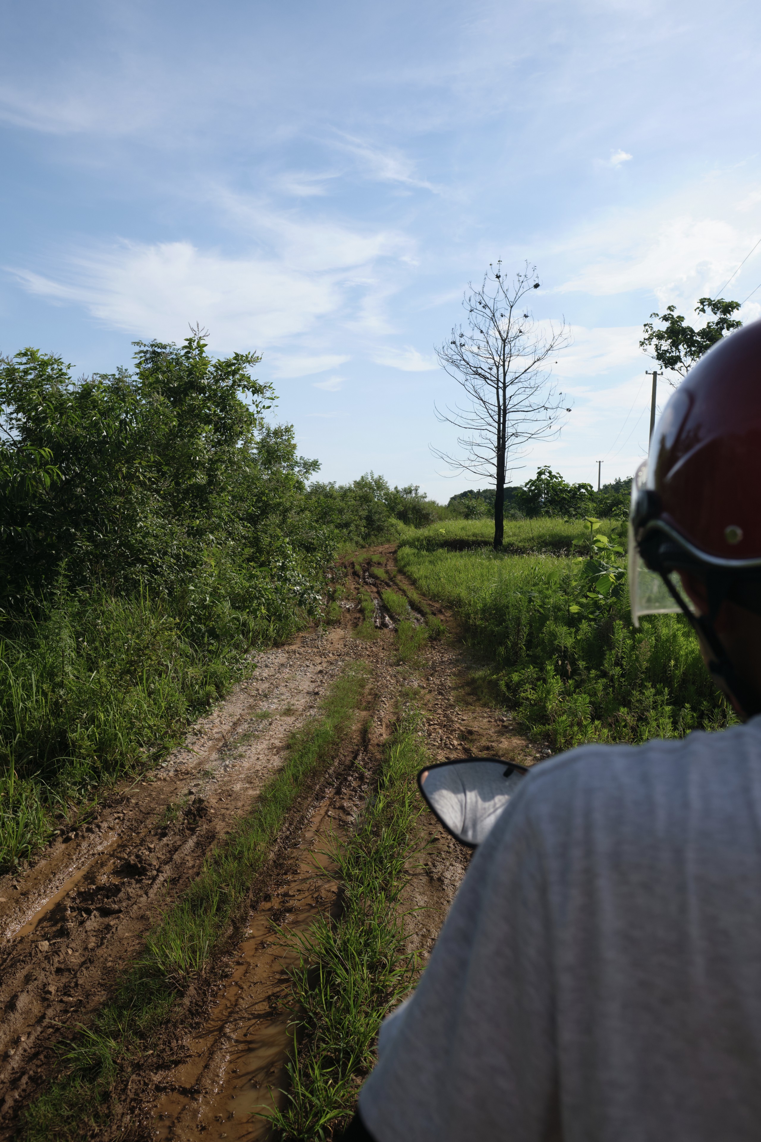 View from behind a person wearing a red helmet on a muddy dirt path with grass and trees under a partly cloudy sky