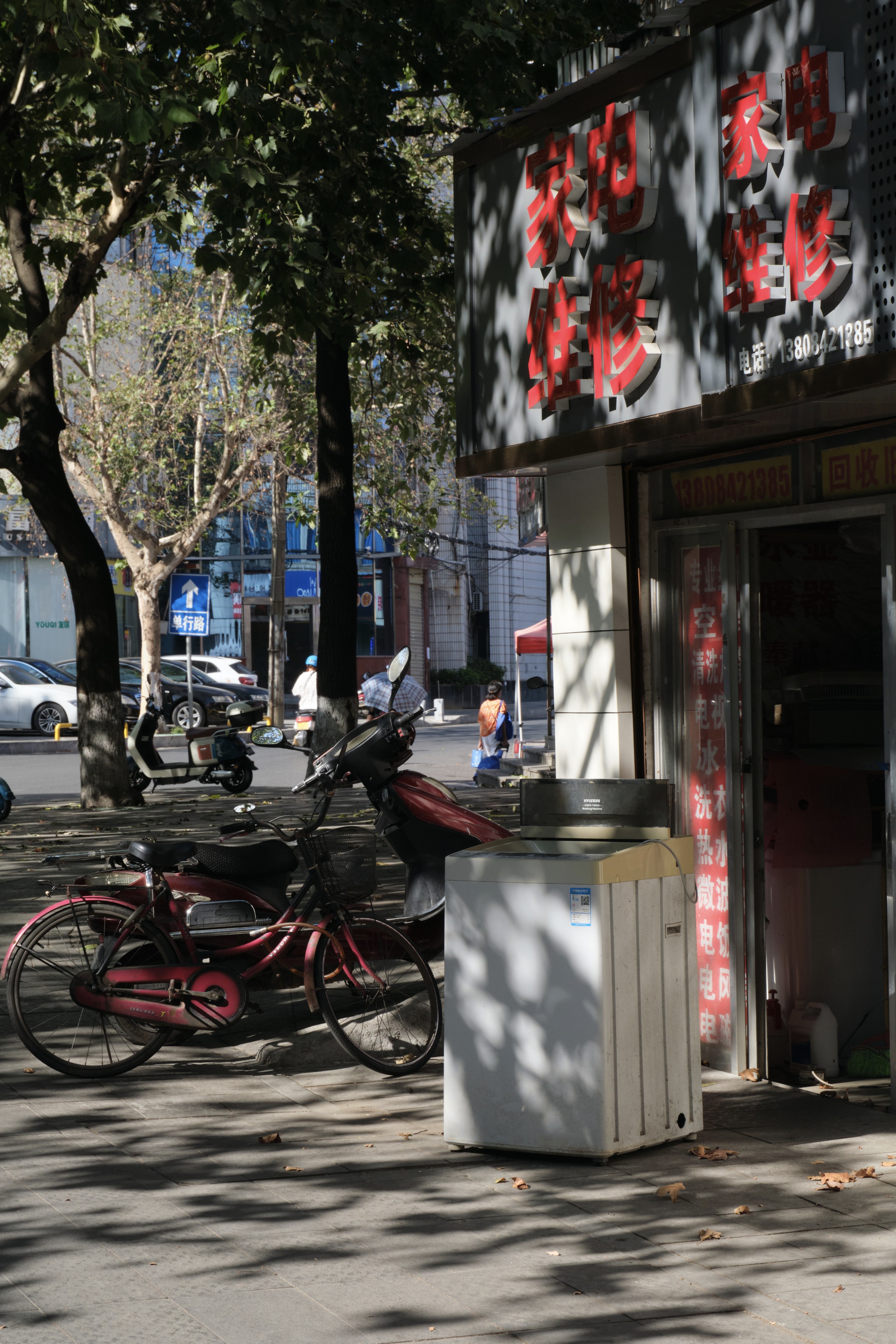 Red bicycle and white refrigerator on a sunlit sidewalk in front of a building with red Chinese characters. Trees line the street with parked cars and blurred pedestrians in the background