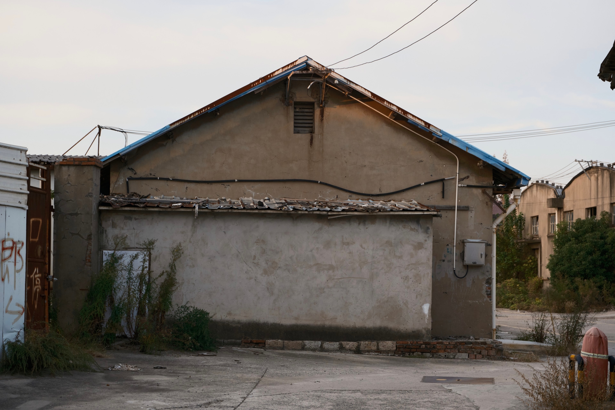 Aged concrete building with gabled roof, visible wires, a small high window, and a low stucco wall in front