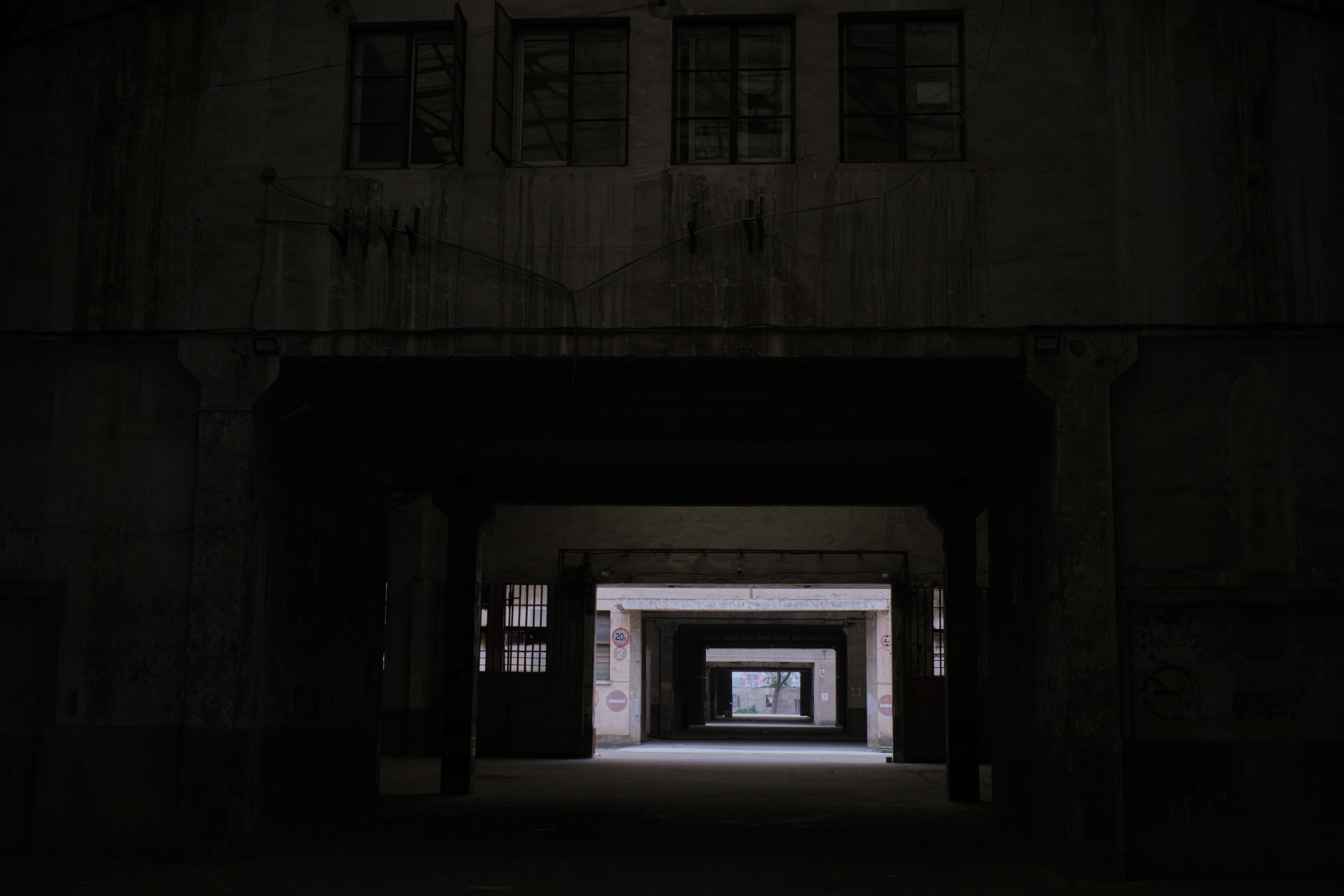 View through a dark, long building passage revealing multiple doorways and a brightly lit distant exit. Overhanging structure with windows