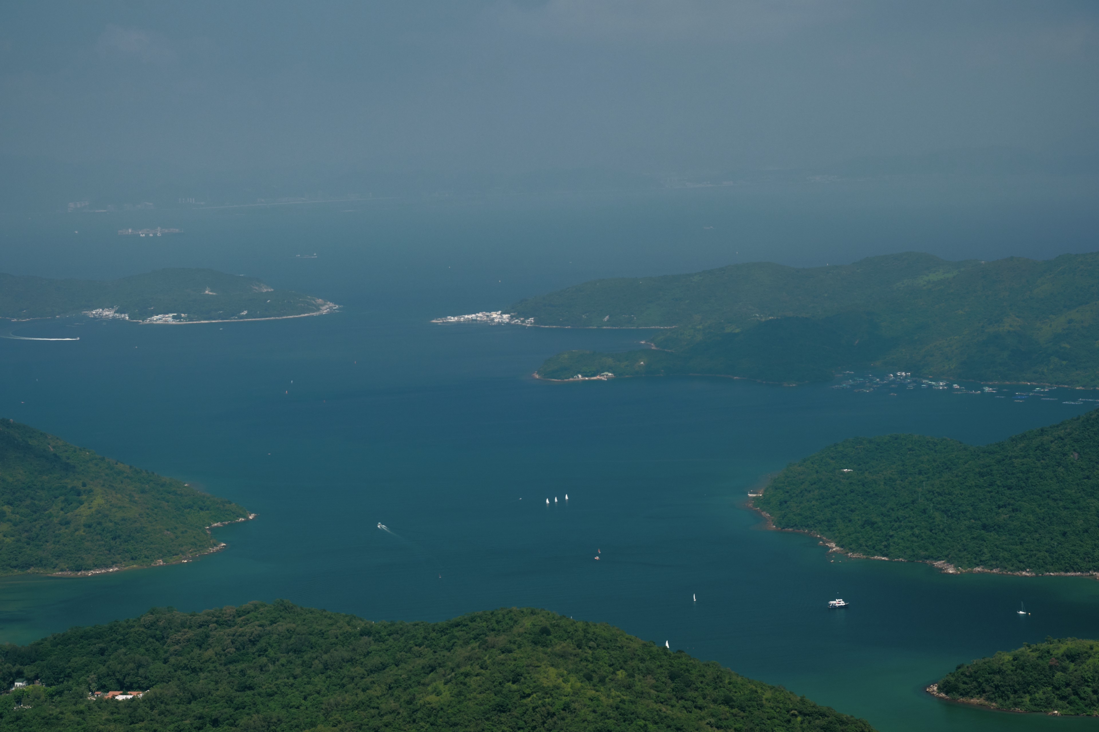 Aerial view of a blue-green sea inlet flanked by lush green, forested islands with multiple white boats, under a hazy sky