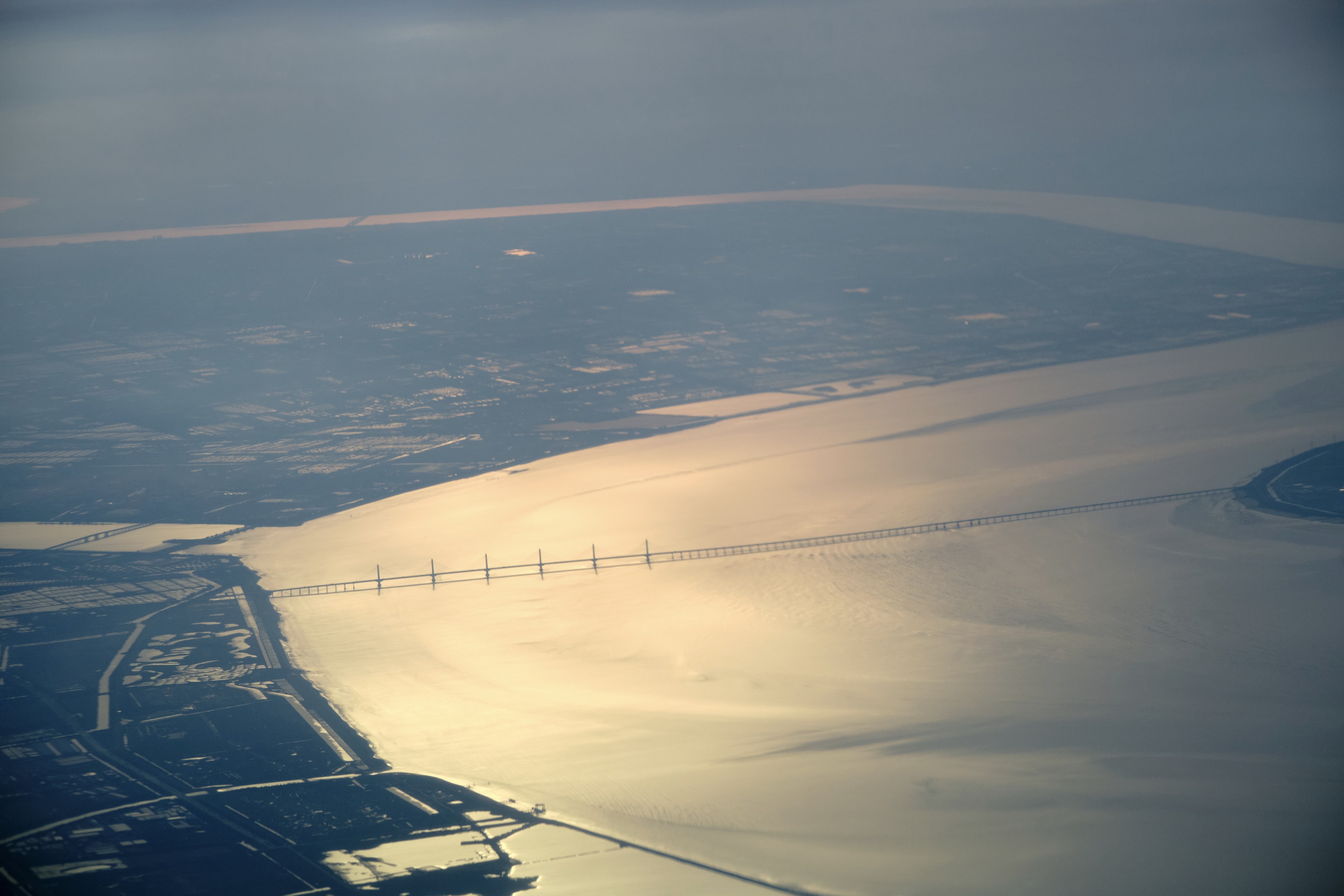 Aerial view of a long bridge spanning a wide, sun-dappled body of water, with hazy landmasses on both sides