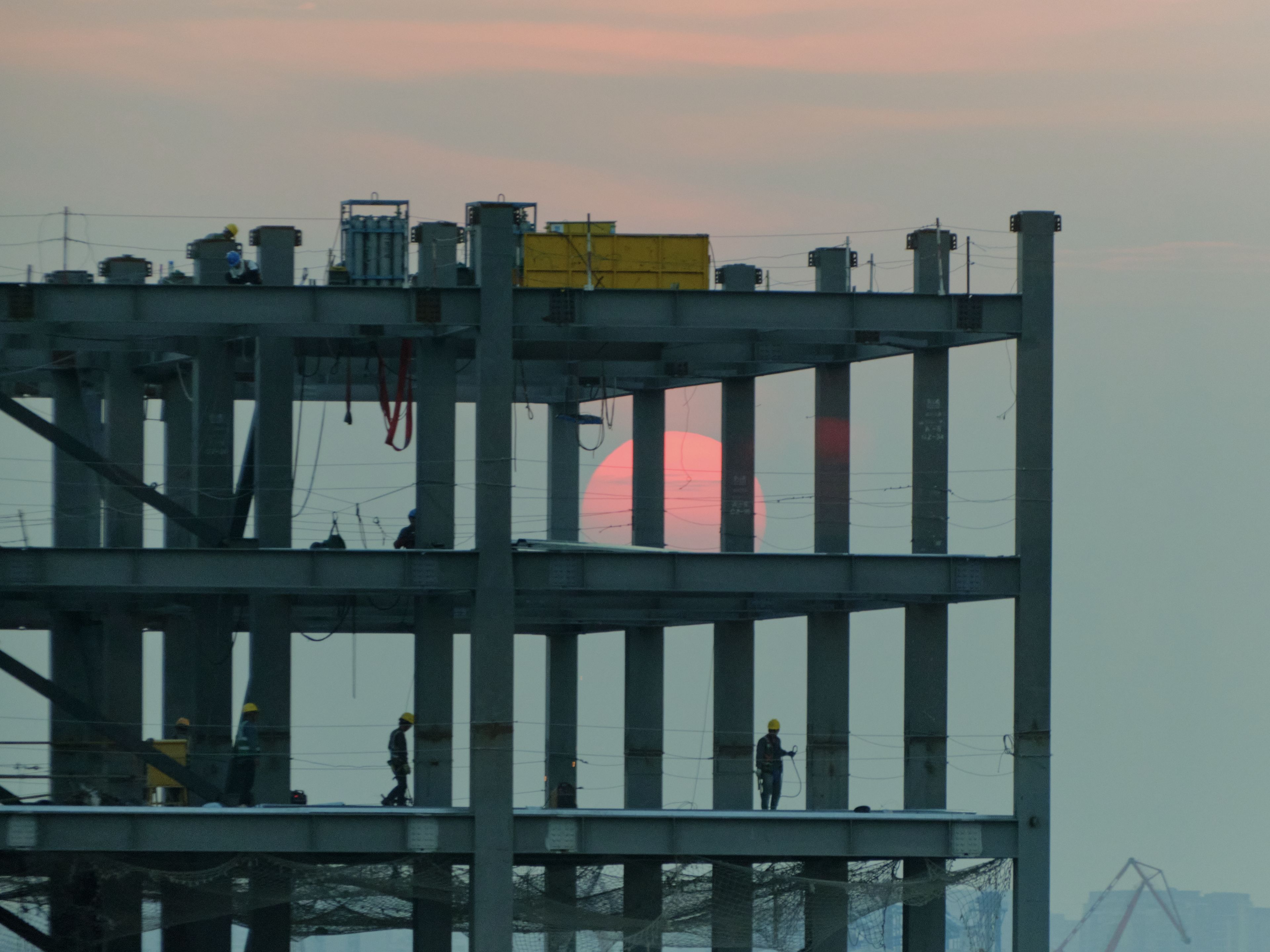 Steel framework of a multi-story building under construction with a large red sun visible through its upper levels. Silhouetted workers are on the lower floors against a pale sky