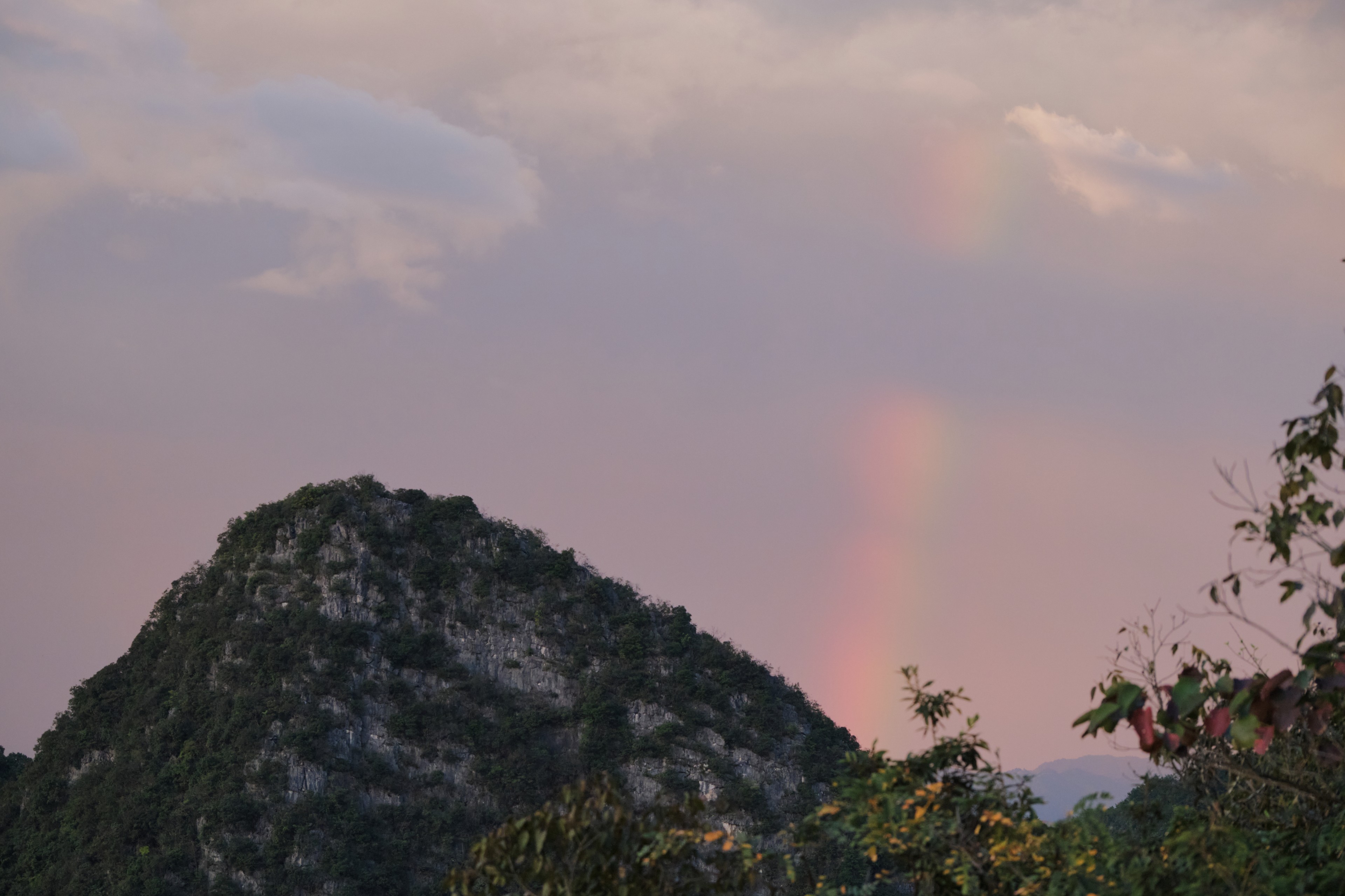 Dark forested mountain peak under a light pink sky with a faint vertical rainbow effect and sparse clouds, with foliage visible on the right