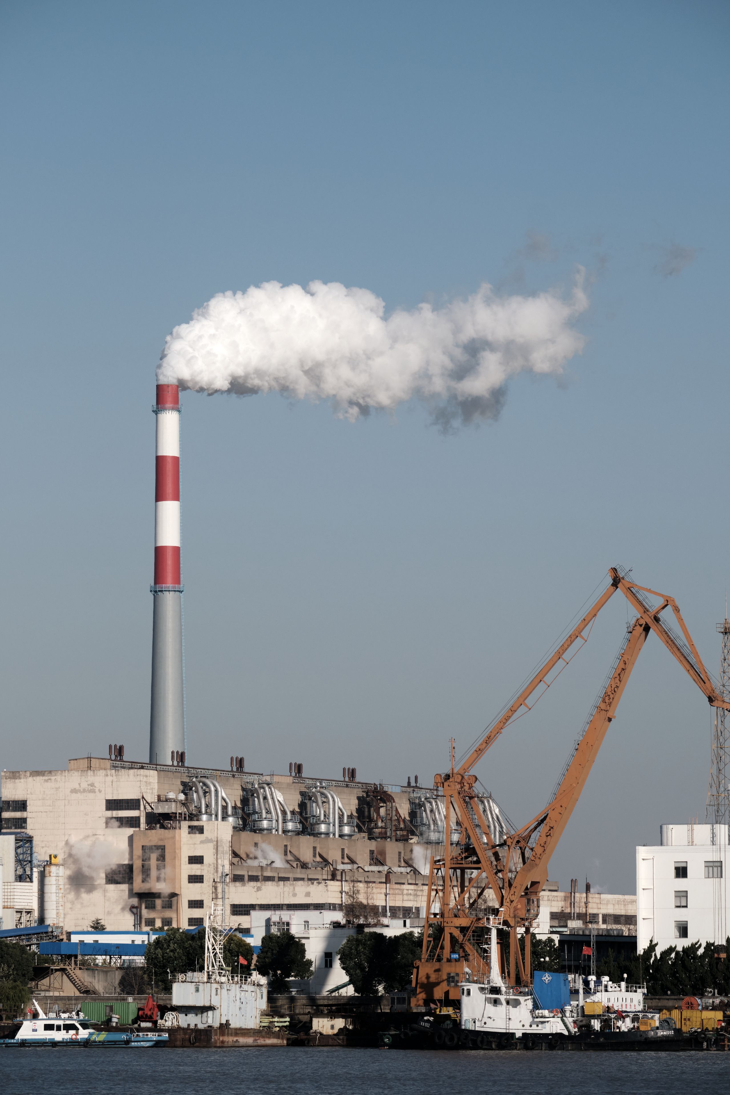 Smokestack emitting white smoke above an industrial building, with orange cranes and water in the foreground under a blue sky
