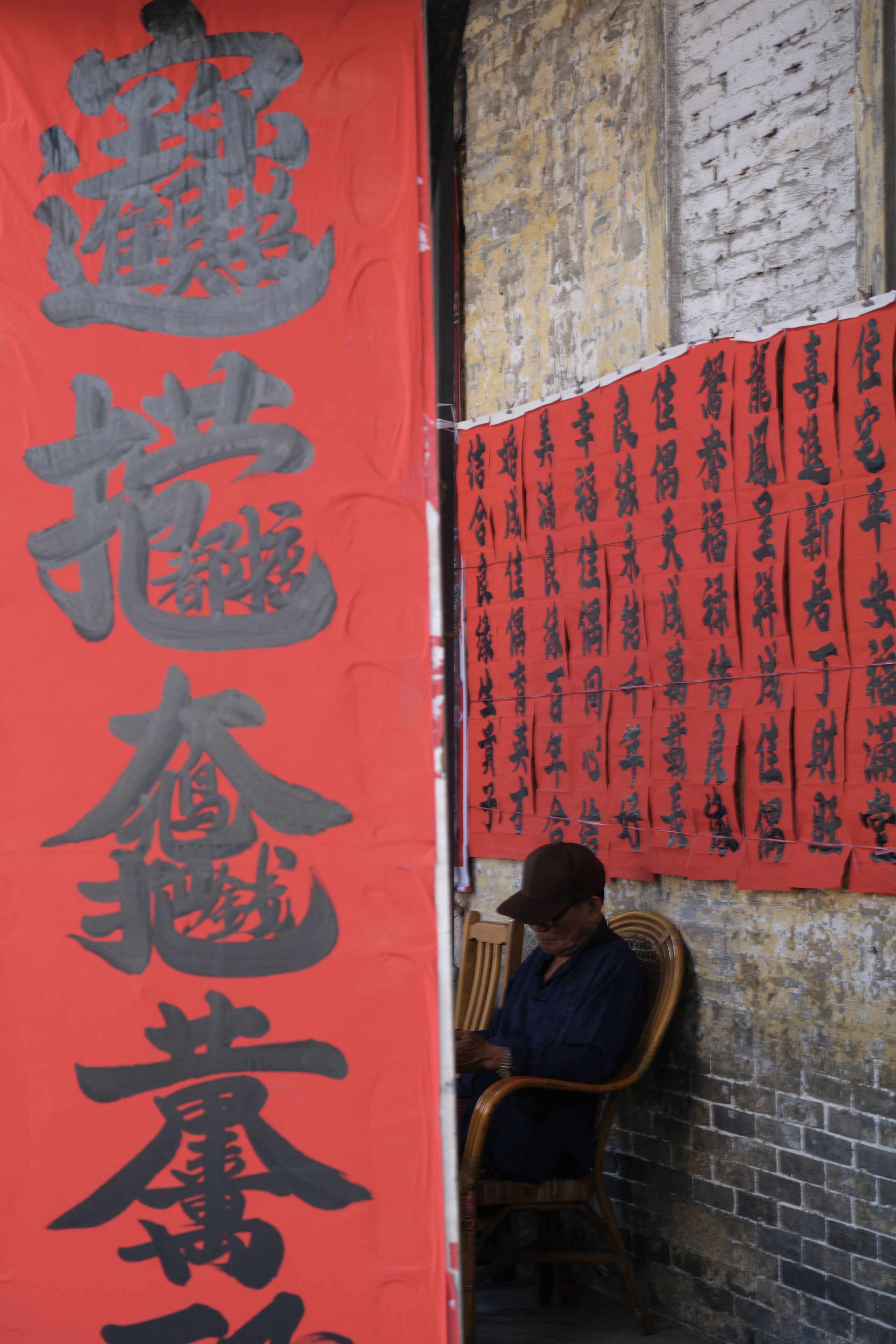 A man sits on a wooden chair against a brick wall, partially obscured by a large red banner with black Chinese calligraphy. Smaller red banners with calligraphy hang on the wall to the right