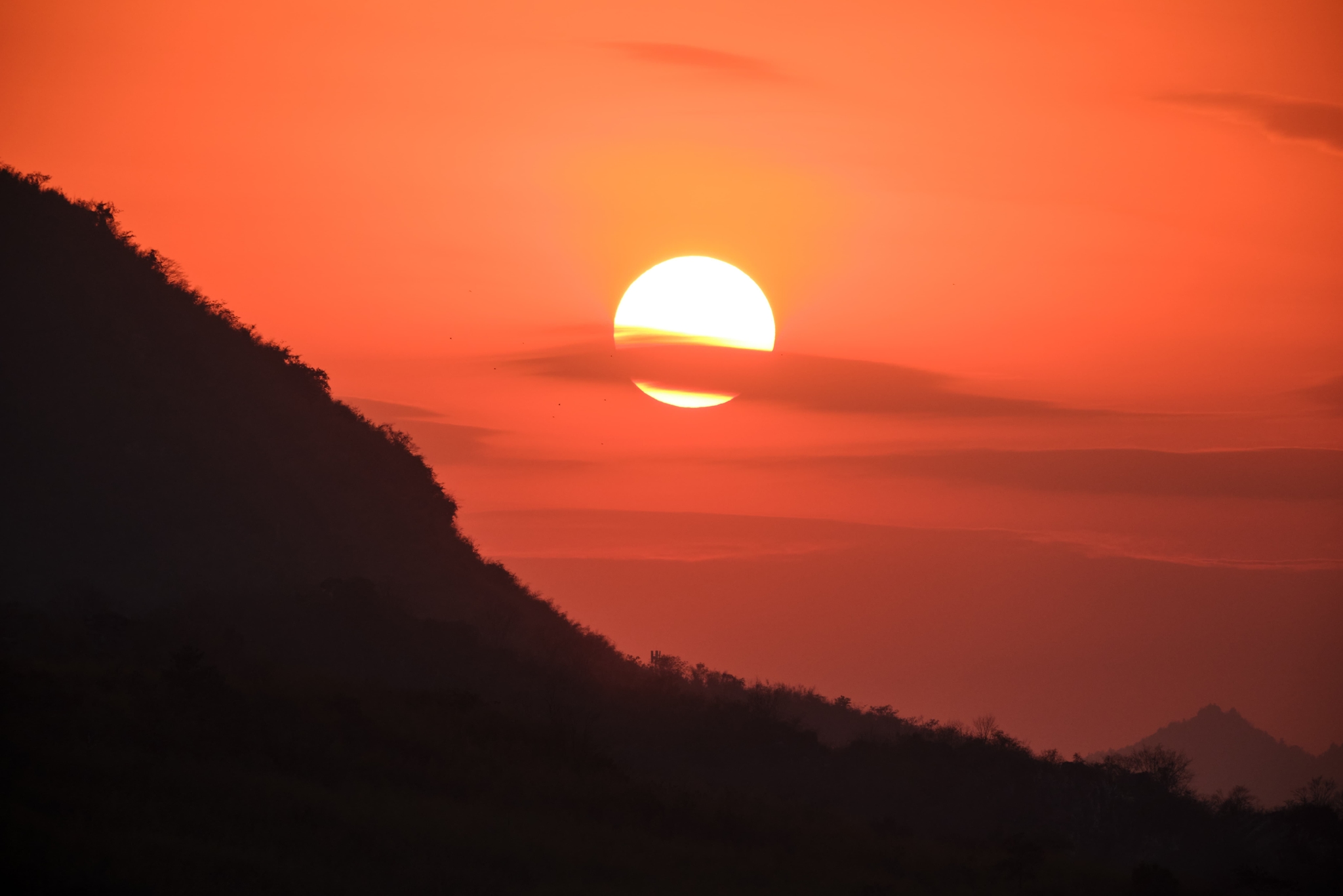Bright sun centered against an orange and red sky, with a dark silhouetted mountain slope occupying the left foreground and subtle horizontal cloud bands