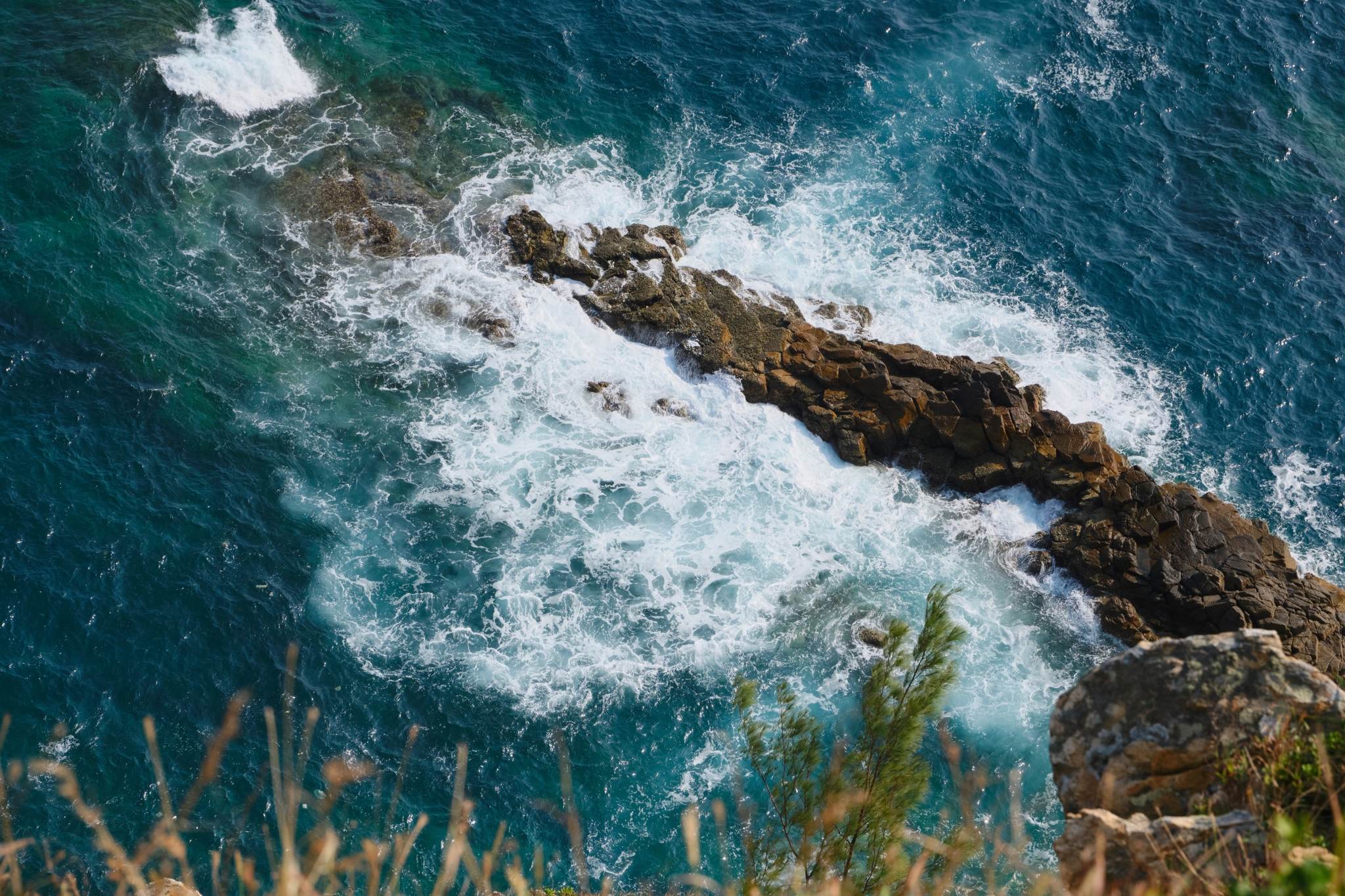 Aerial view of waves breaking against dark rock formations in deep blue and turquoise ocean water, with blurred foreground vegetation
