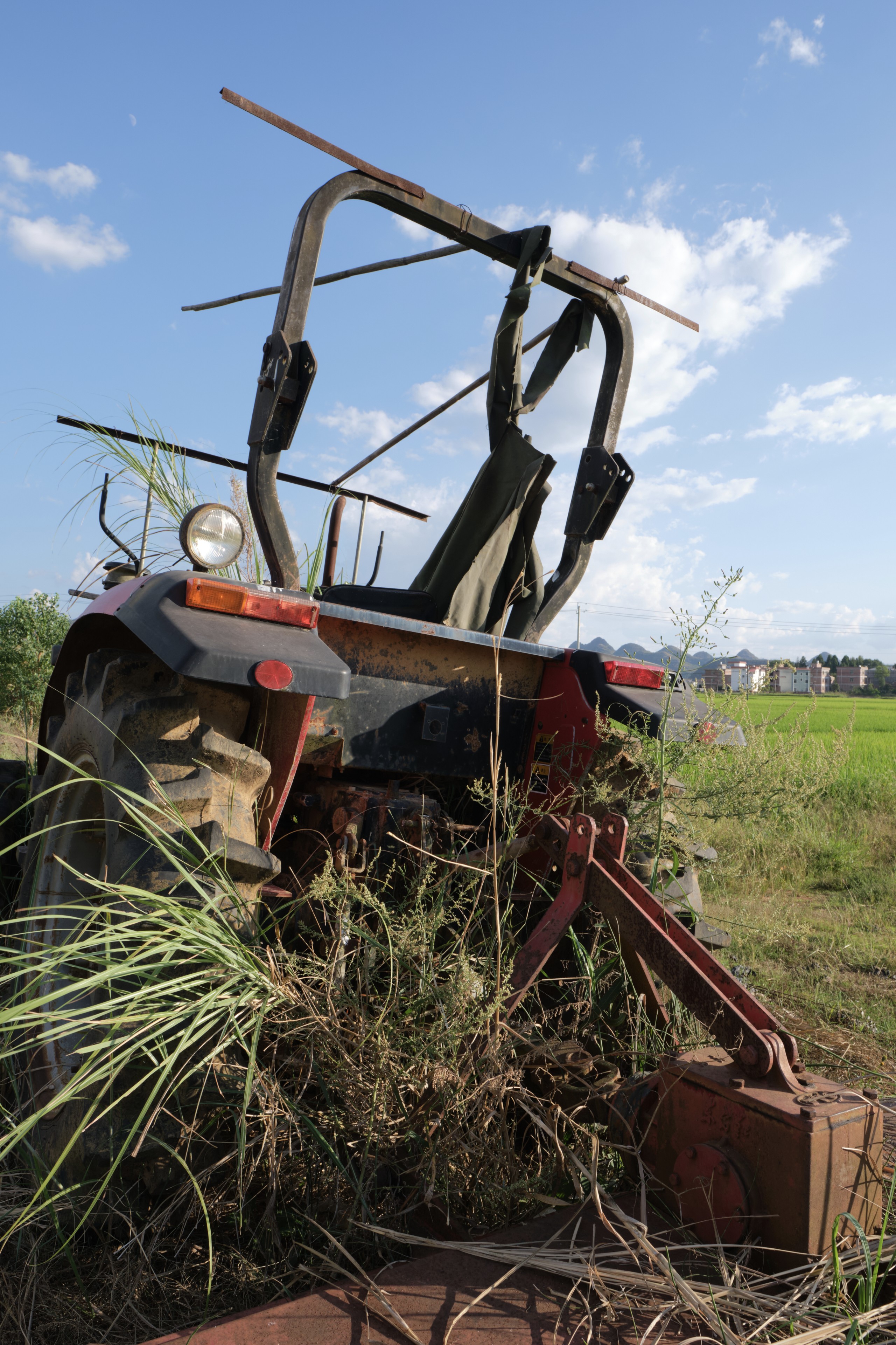 Rear view of a red and black tractor partially obscured by overgrown green grass in a field. A bright blue sky with sparse clouds is above, and distant buildings are visible on the horizon
