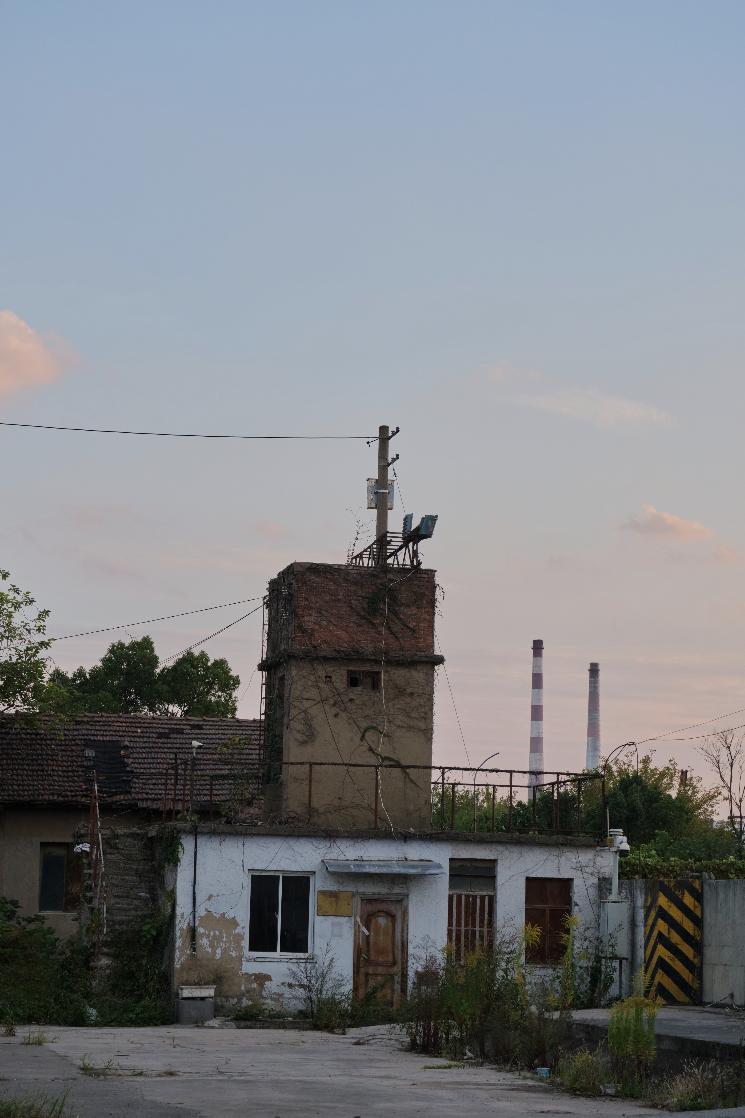 A cluster of dilapidated buildings with a central tower and industrial smokestacks in the background under an evening sky