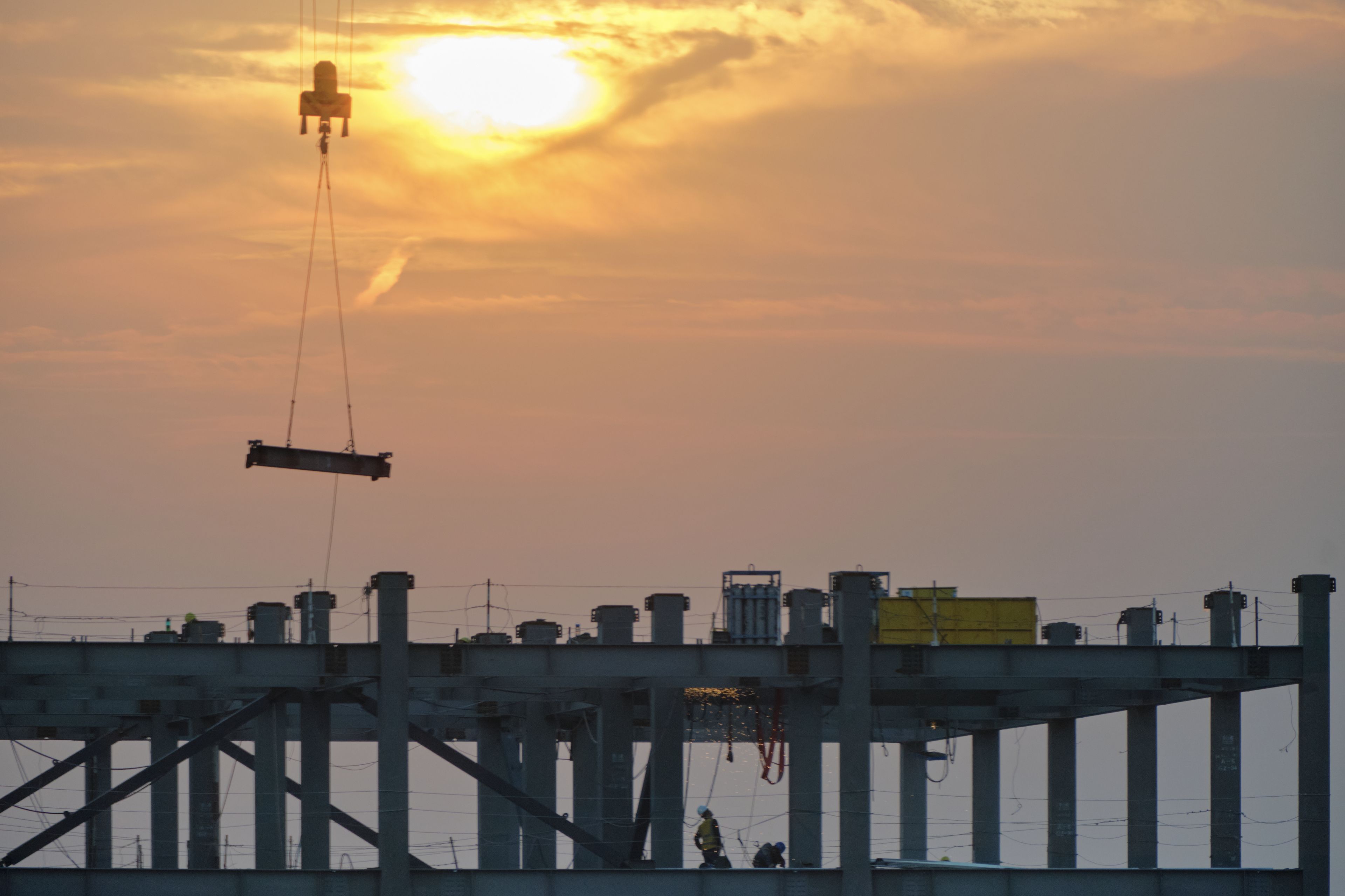 Orange sky with setting sun above silhouetted construction site. Crane lowers long object over concrete pillars and beams. Faint contrail present