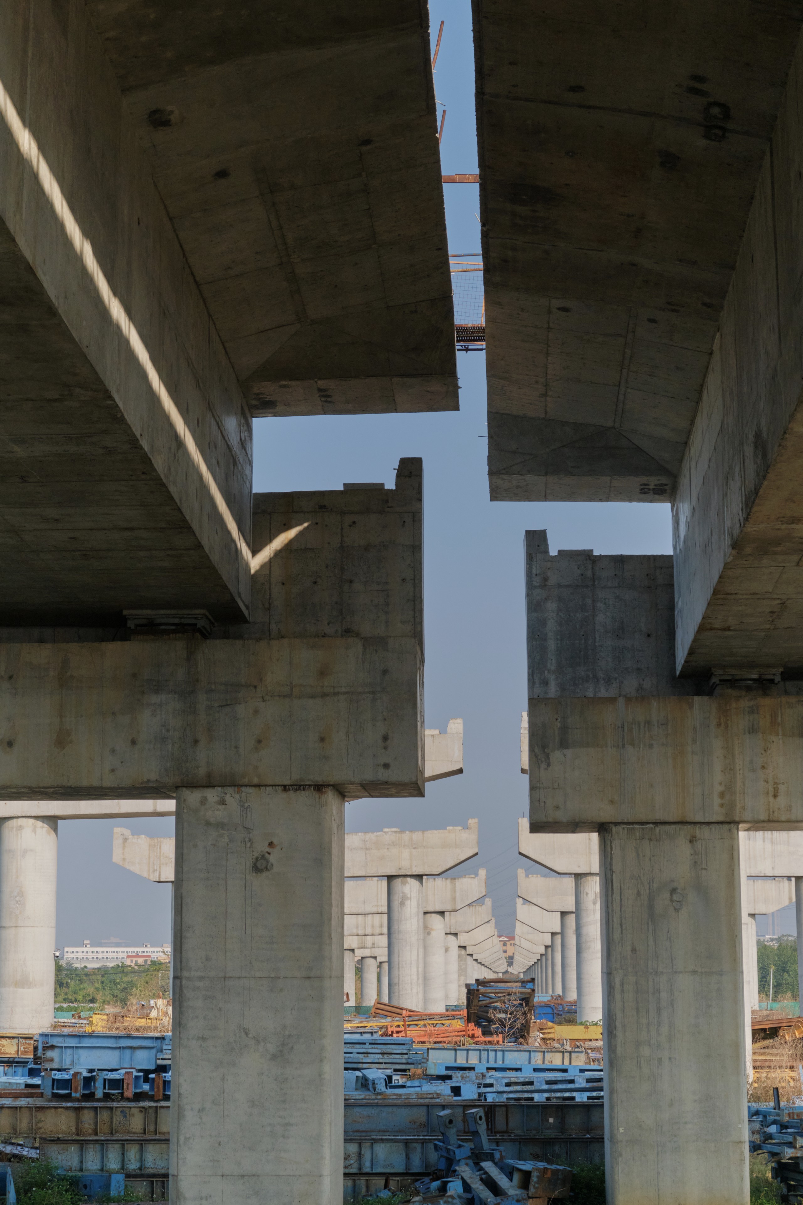 View from beneath a massive concrete elevated structure under construction, revealing multiple geometric pillars and beams against a clear sky. Distant unfinished sections of the structure are visible, with sunlight highlighting concrete surfaces