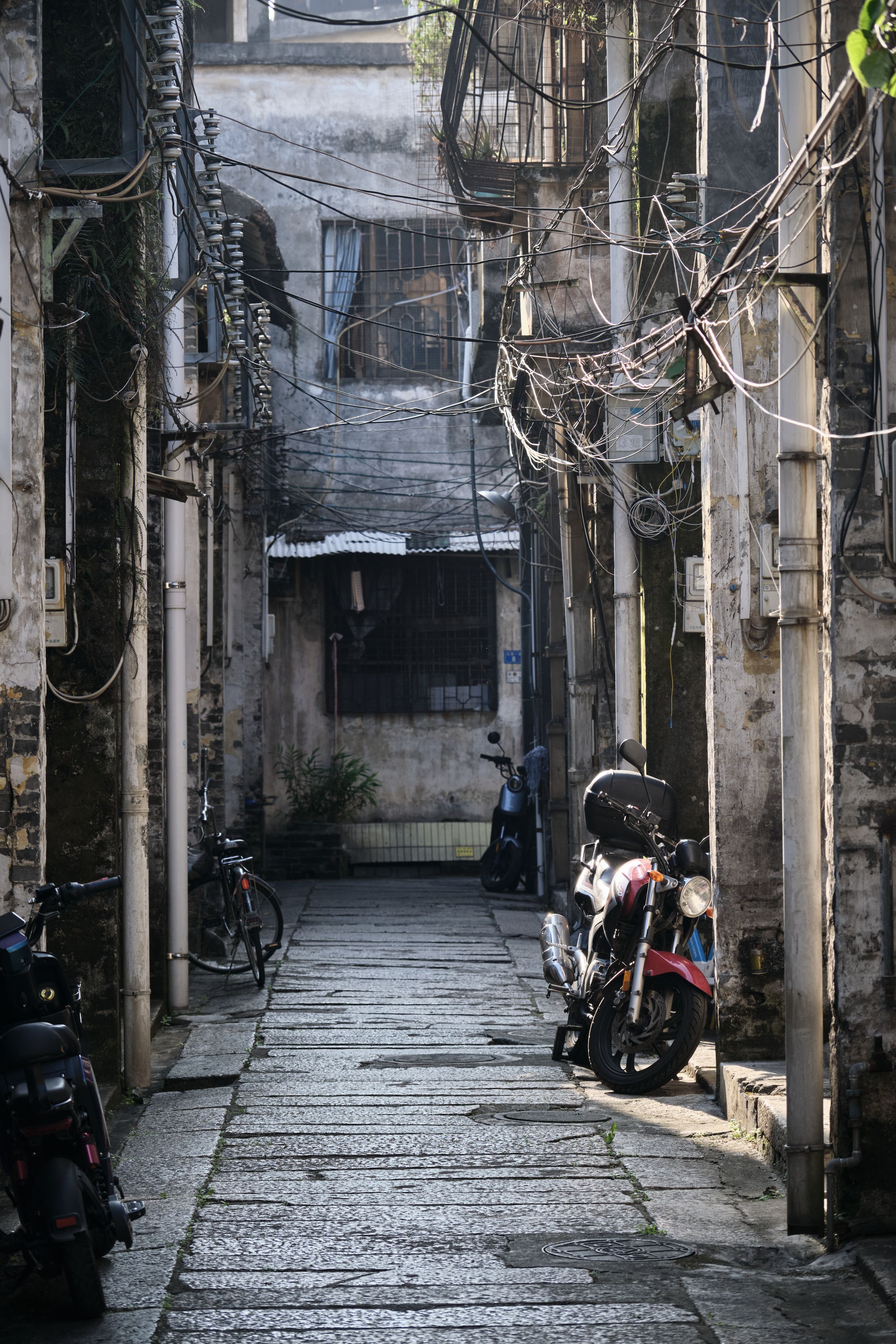 Narrow, vertical view of a densely wired alleyway with parked motorcycles and bicycles between old buildings