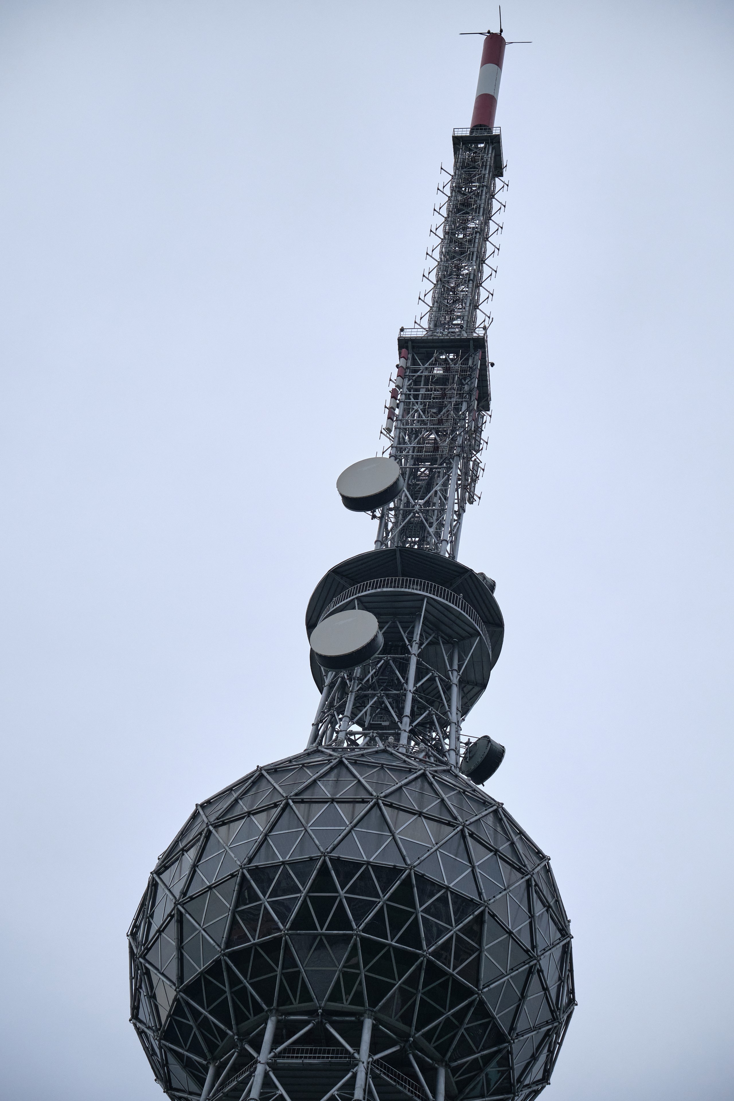 Close-up of a tall steel transmission tower's spherical observation deck and an antenna mast extending against an overcast sky
