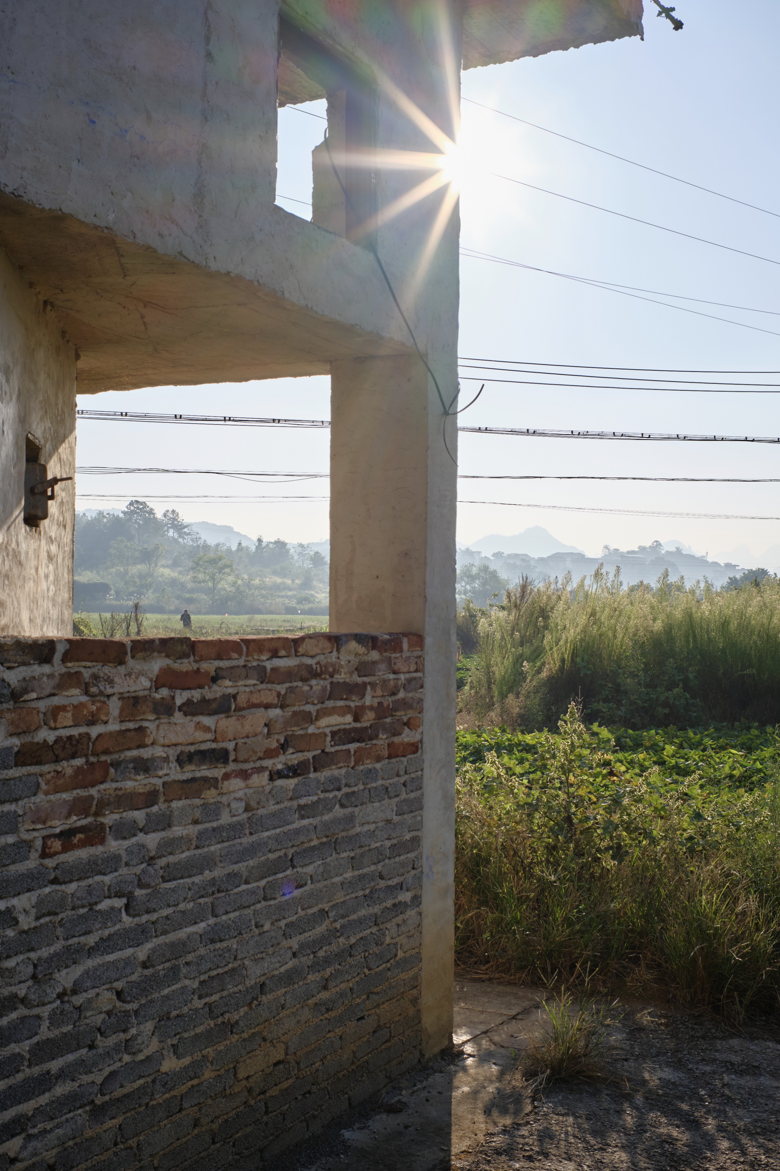 Sun flares from a concrete structure with an exposed brick wall, overlooking green fields and distant hills under a clear sky with power lines
