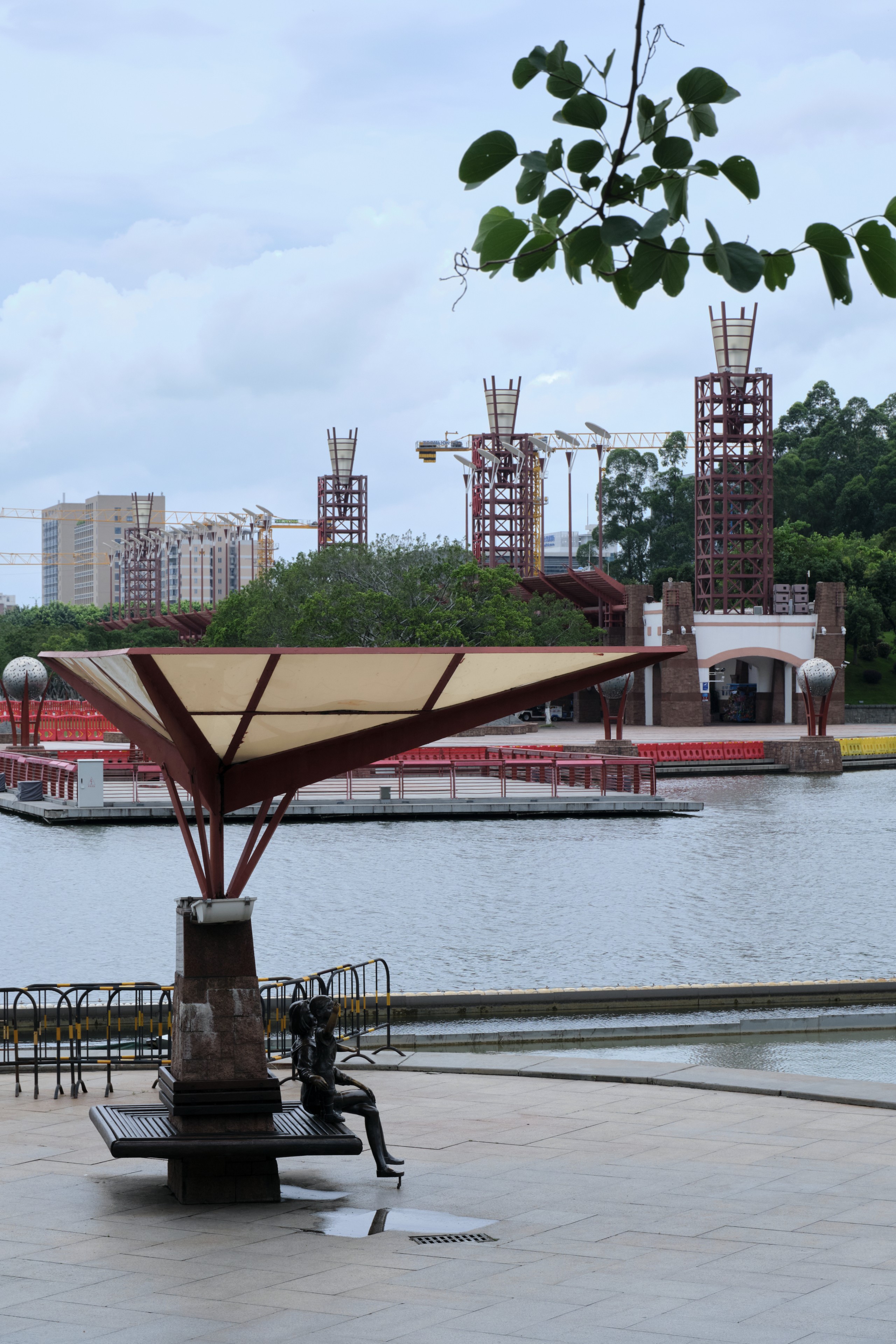 A modern park area with a statue on a bench under a stylized sunshade, facing a large body of water. Across the water, several tall, sculptural structures are under construction, with residential buildings and trees in the background under an overcast sky
