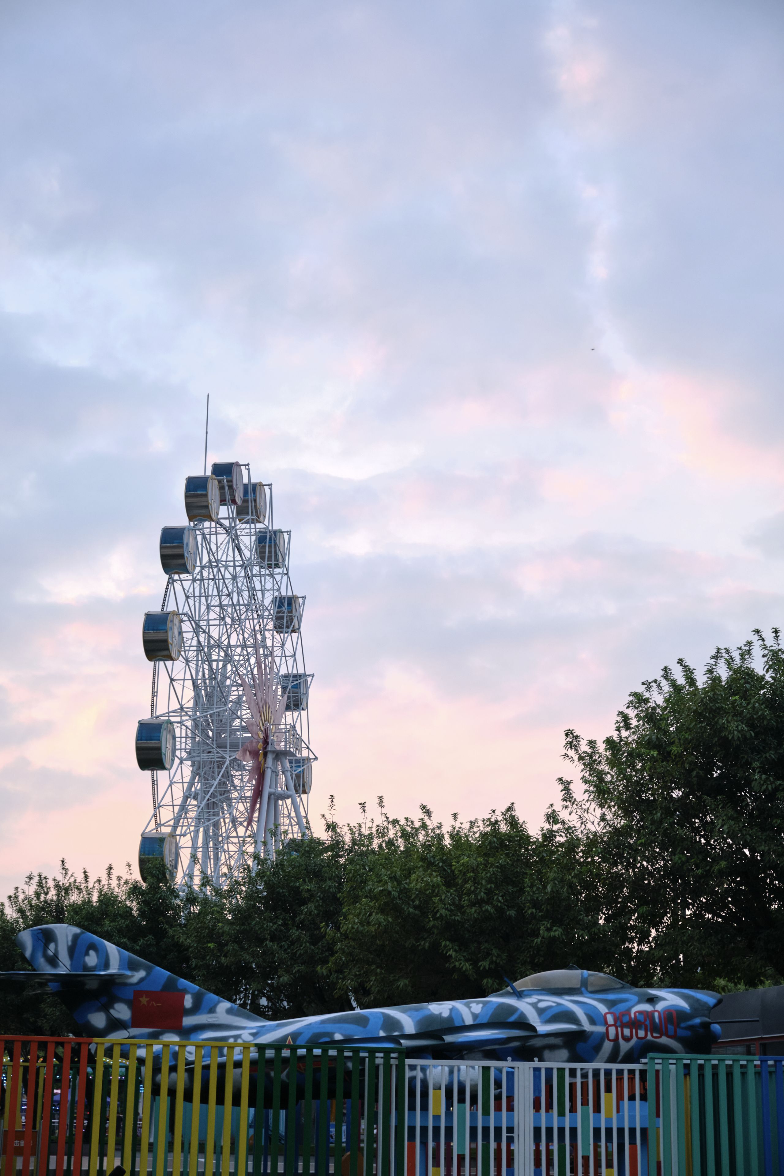 White Ferris wheel against cloudy pink-blue sky, airplane model, green trees, colorful fence