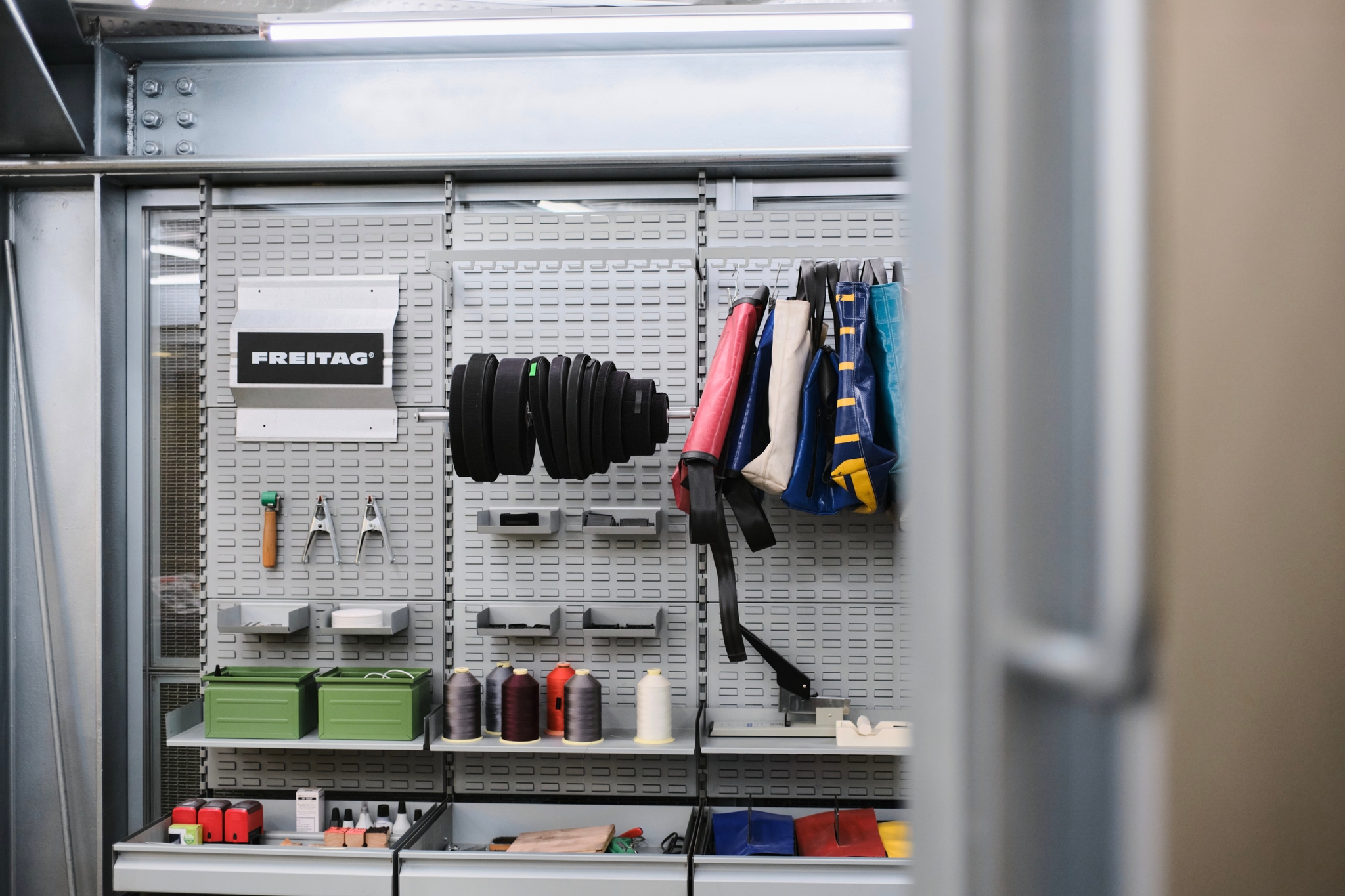 Grey pegboard wall displaying tools, spools, colorful fabric bags, and a 'FREITAG' branded box in an industrial setting