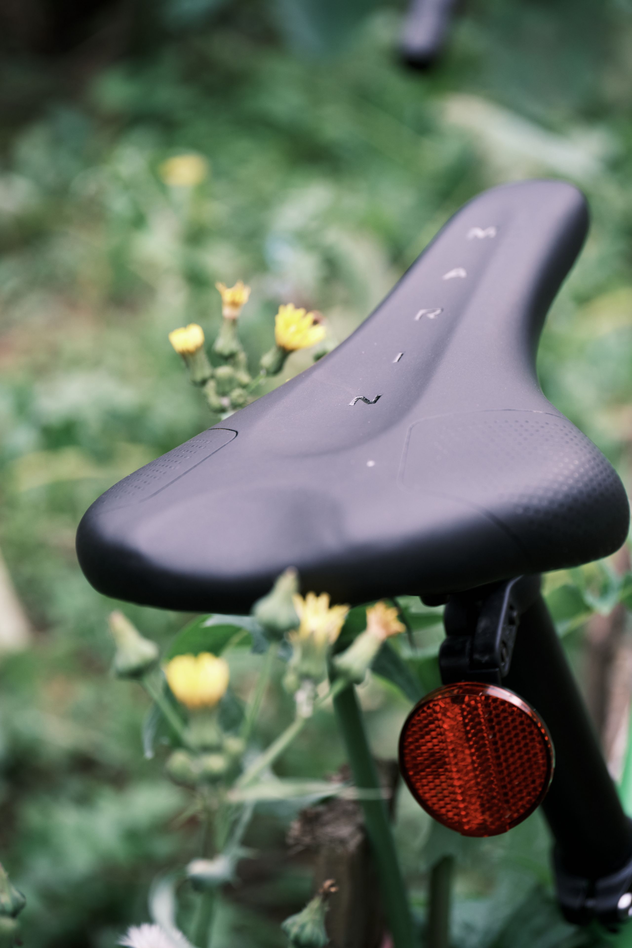 Black bicycle seat with water droplets and red reflector, against blurred green foliage and yellow flowers
