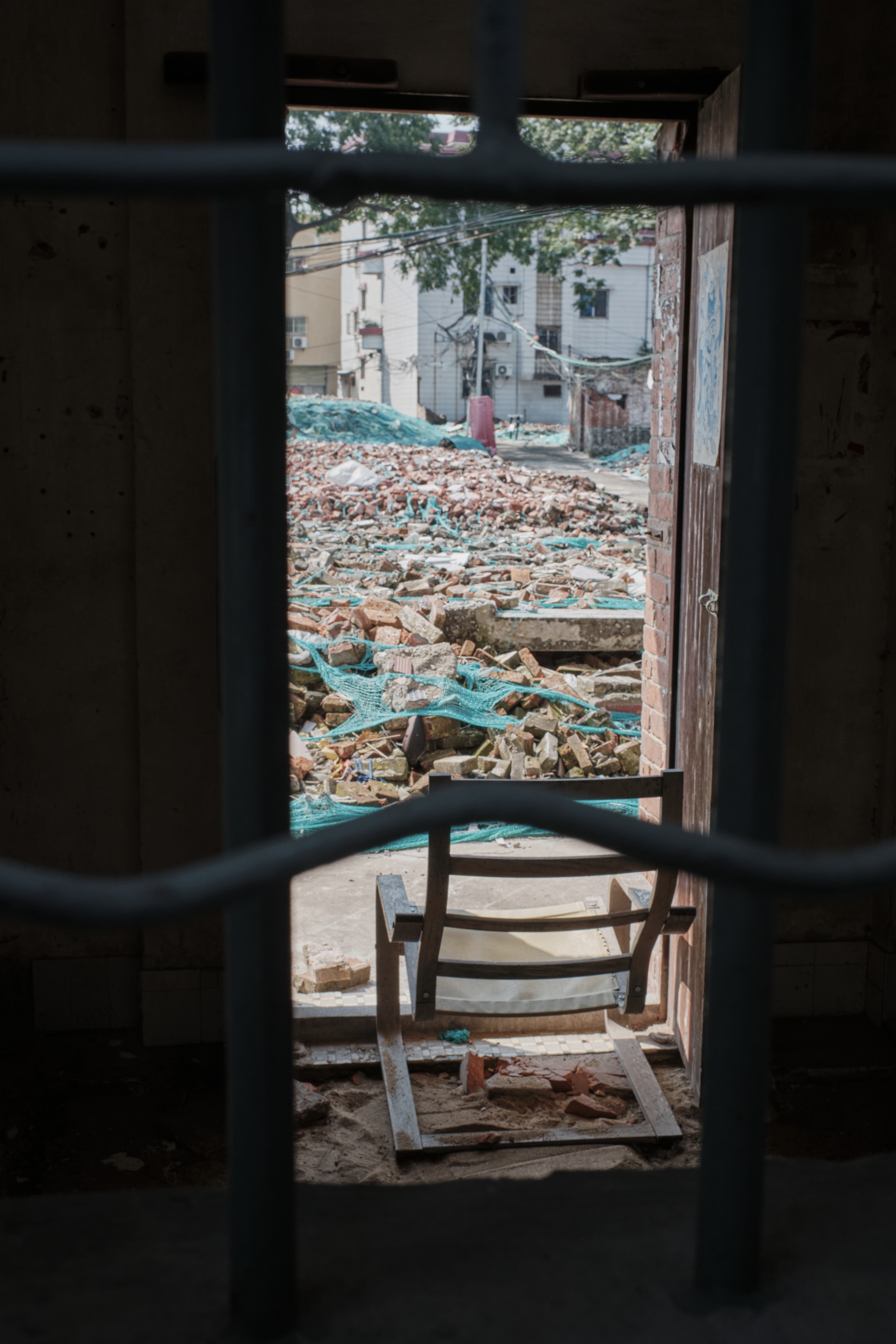 View through dark metal bars and an open doorway revealing an outdoor scene of extensive rubble, including bricks and blue tarps, beneath distant buildings and trees. A broken wooden chair rests just inside the doorway, and a figure in red is faintly visible amidst the debris