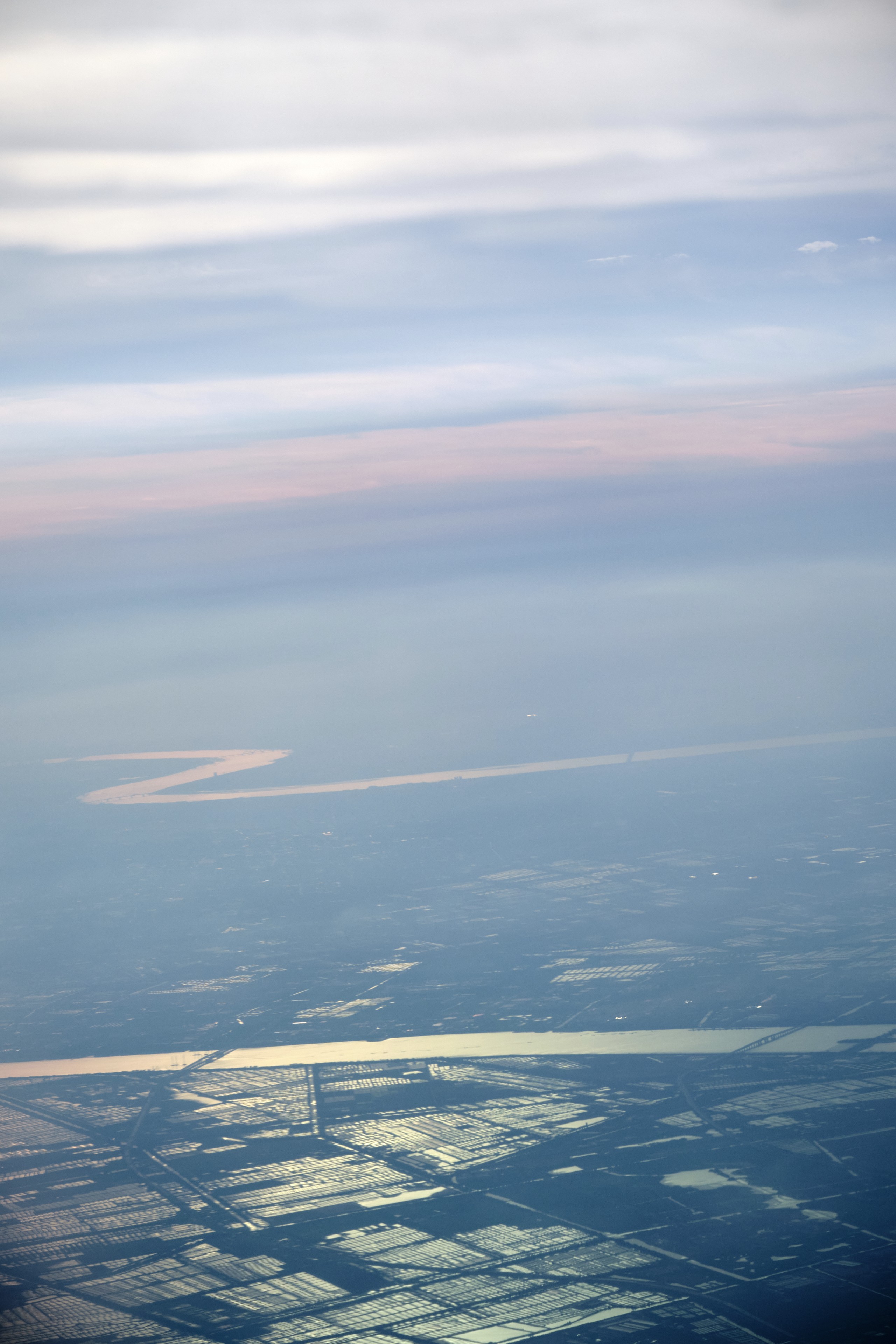 Aerial view of two wide, meandering rivers flowing through a hazy, expansive landscape. The foreground features reflective, geometrically patterned agricultural fields under a soft, layered sky showing light blue and subtle pink hues