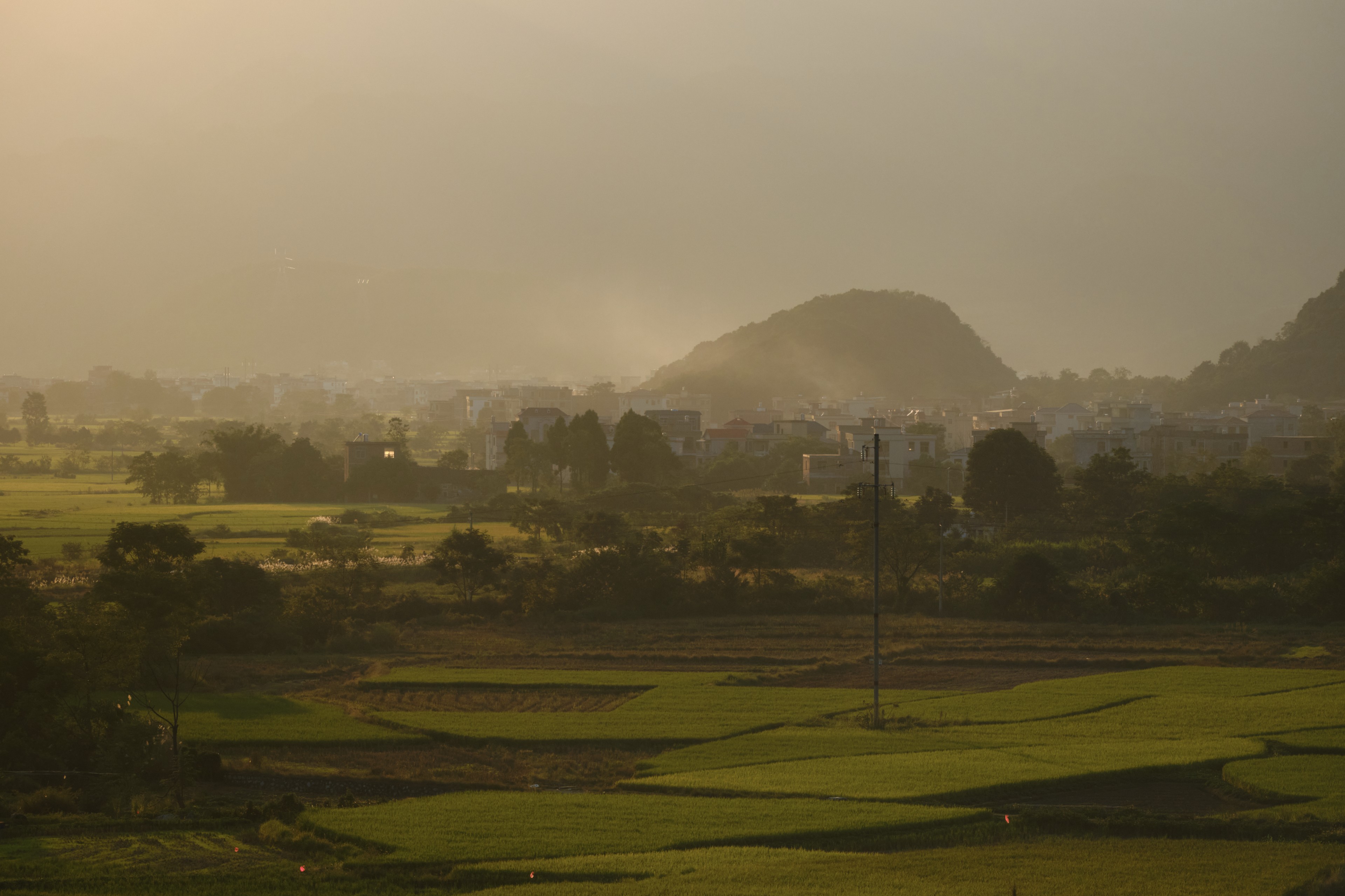 Hazy landscape with green fields, distant town, and hills under golden light