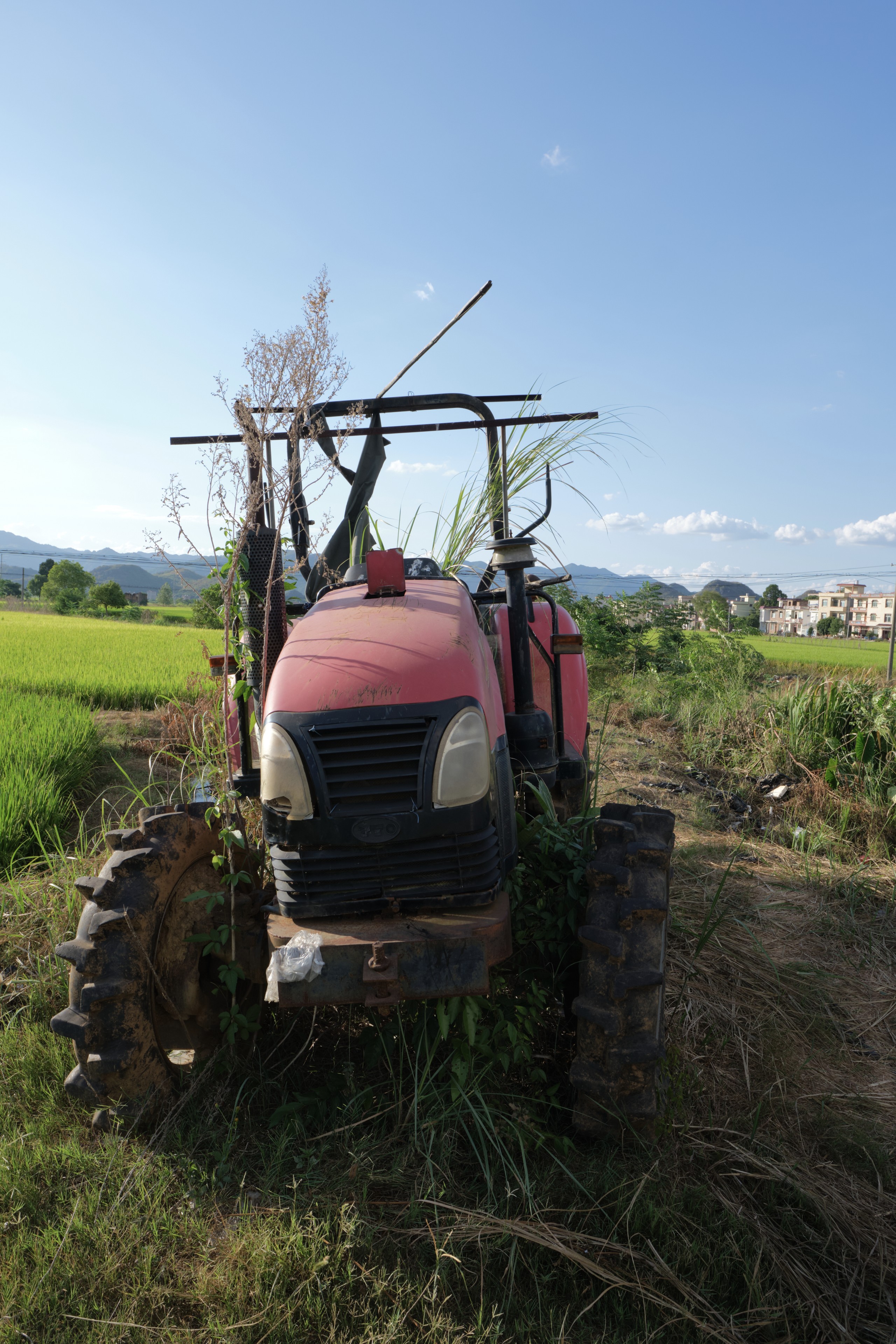 Red tractor overgrown with vegetation in a green field, clear blue sky above, distant structures visible