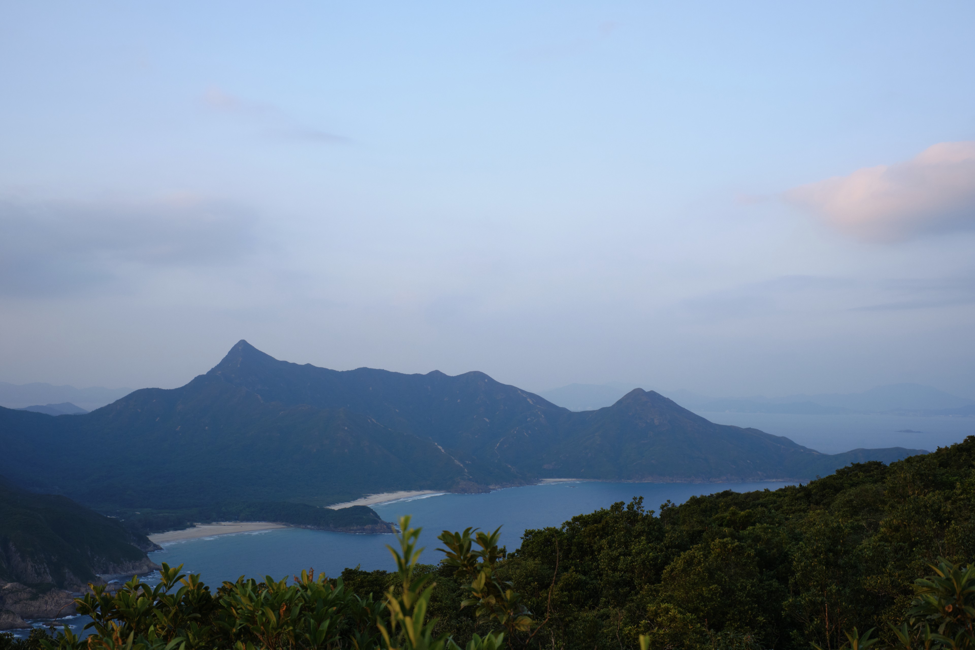 Distant mountains behind a body of water under a light blue sky, with dark green foliage in the foreground