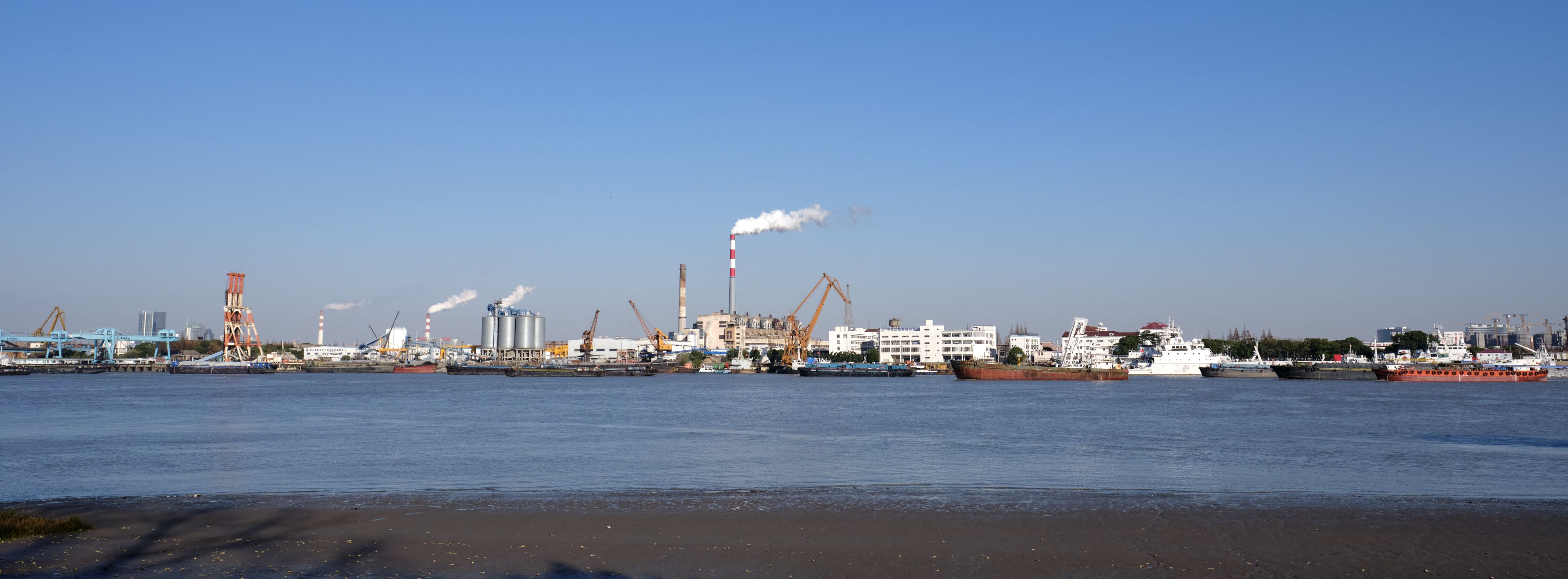 Panoramic view of an industrial port across a body of water with various buildings, cranes, and smokestacks under a clear blue sky. Smoke rises from several stacks. Several vessels are visible along the waterfront