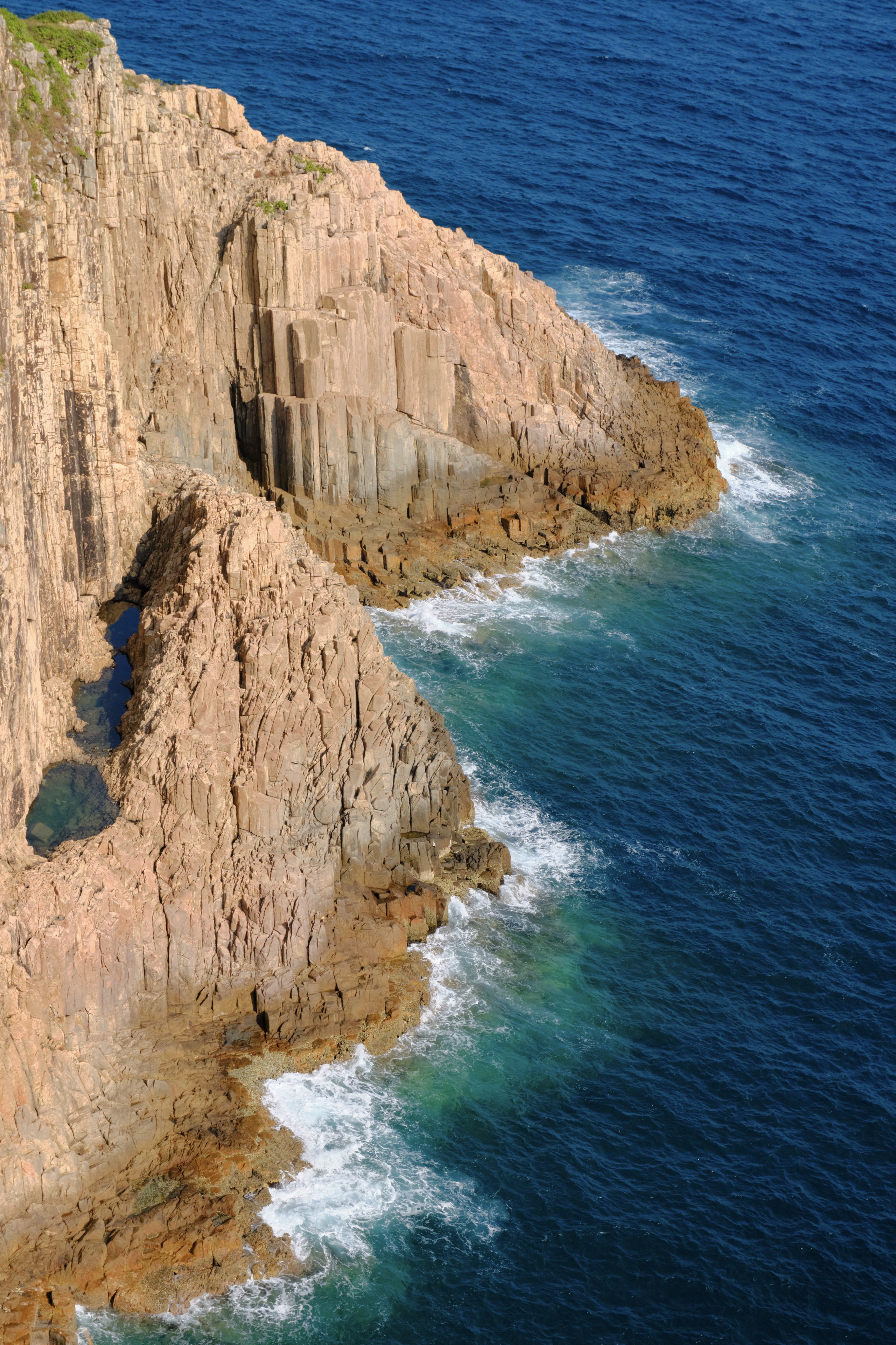 Vertical brown rock cliffs with columnar joints meet deep blue ocean and white waves