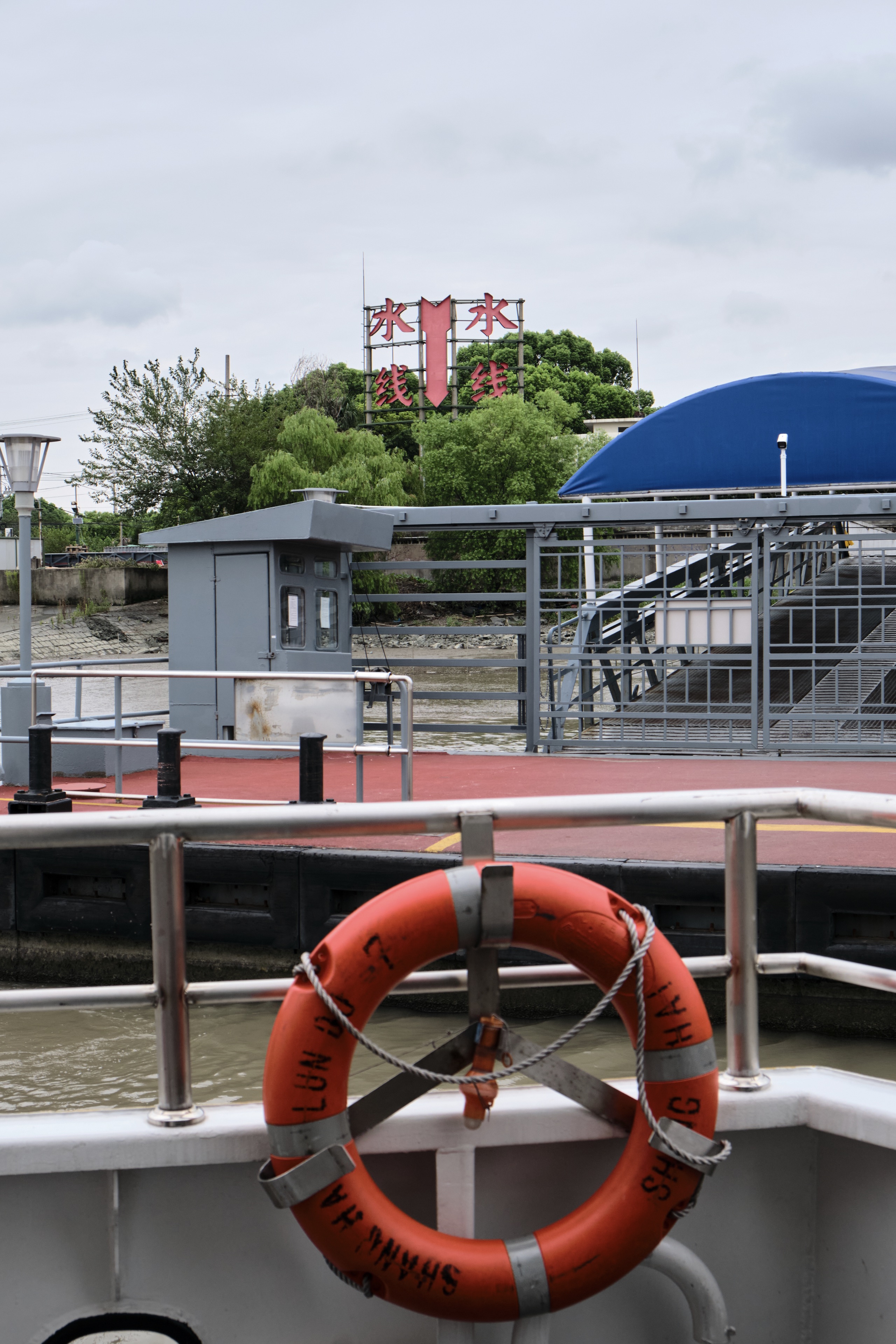 Orange lifebuoy on a white boat railing with a red-decked ferry terminal, gray building, blue canopy, and metal fence behind it. A large red sign with Chinese characters is visible on a distant building amidst green trees under an overcast sky