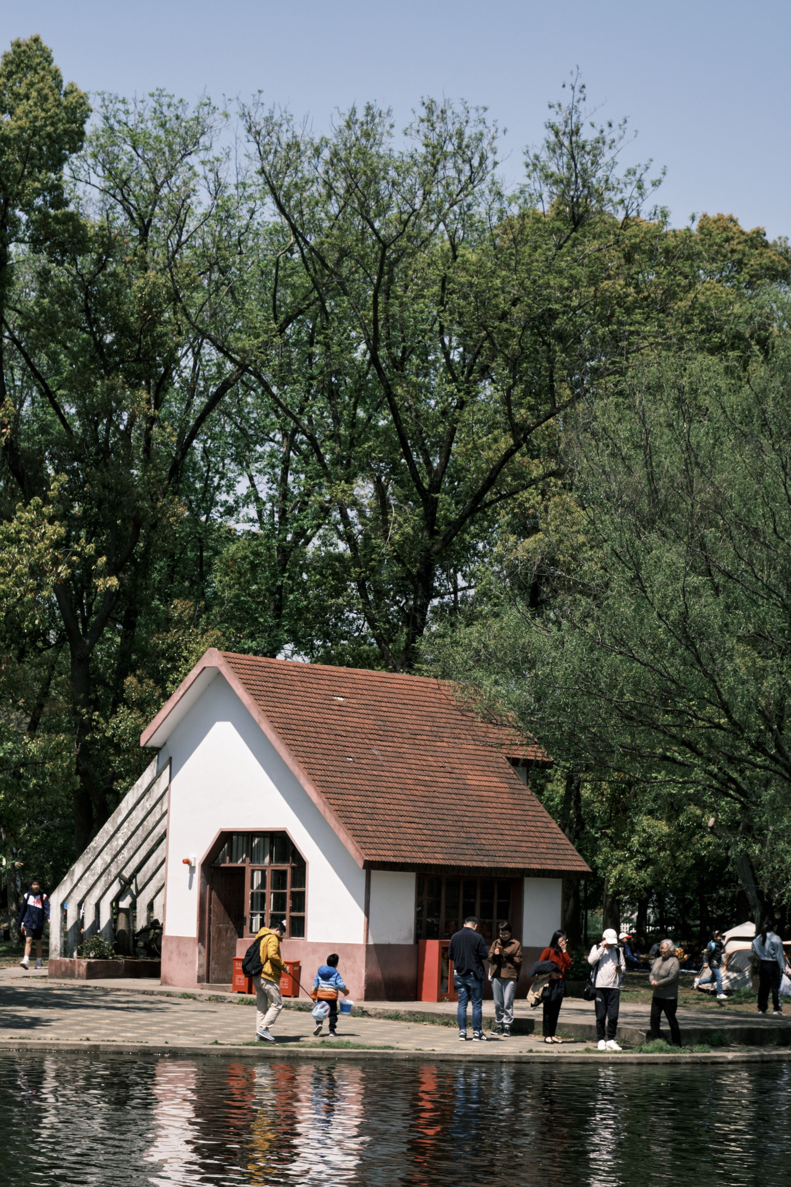 A white building with a red roof stands by a body of water. Several people are visible on the shore and near the building. Lush green trees and a clear sky form the background