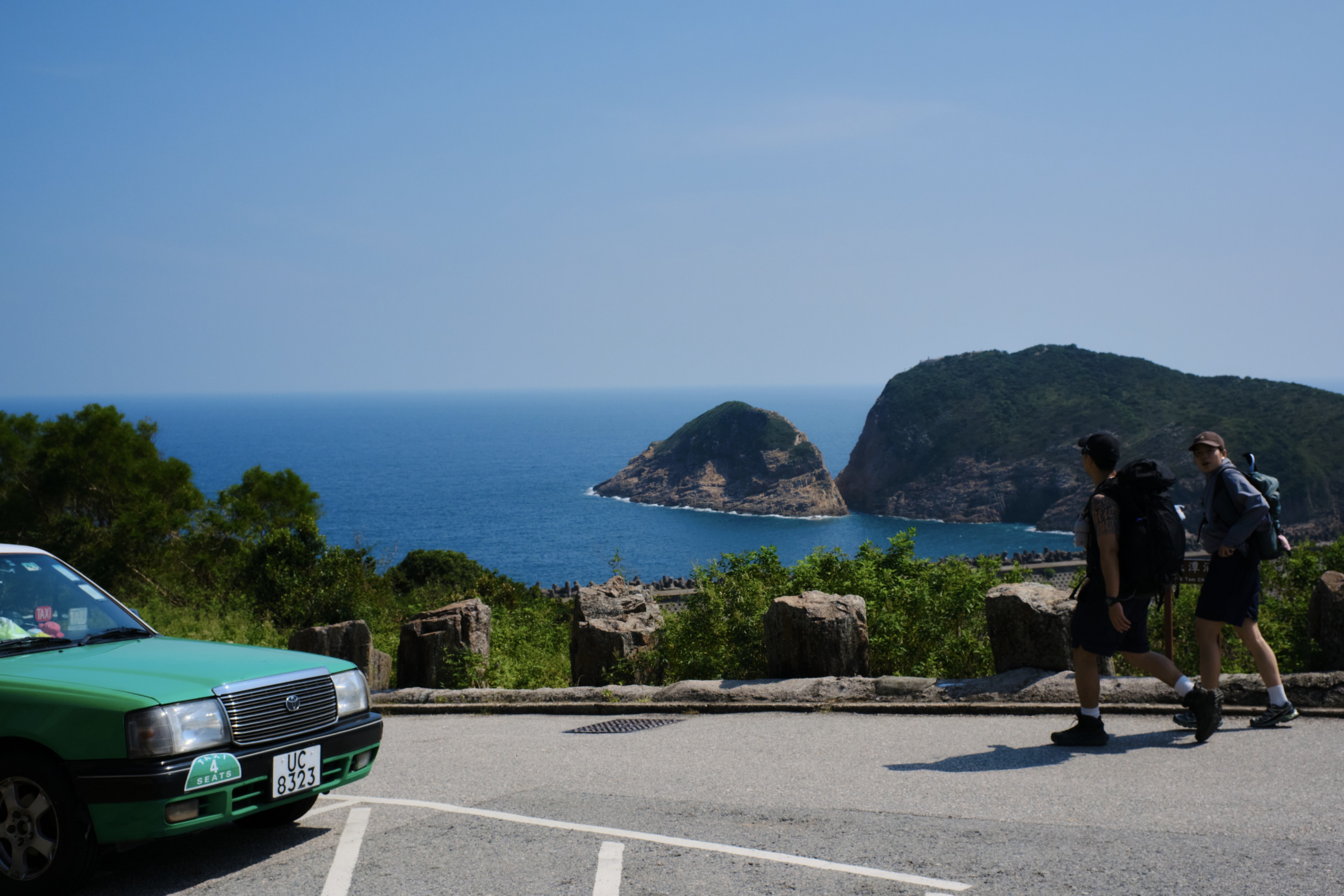Green taxi, two pedestrians, blue ocean, distant islands, coastal road, clear sky