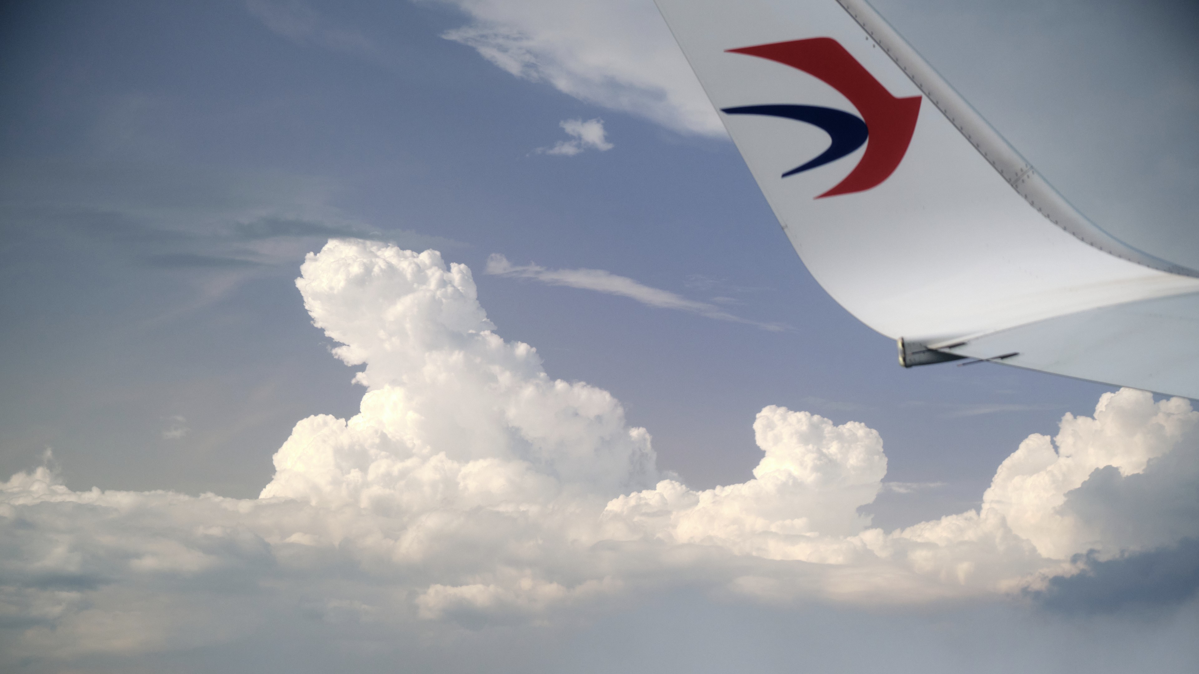 Partial view of an airplane wing with a red and blue logo against a sky with white cumulus clouds