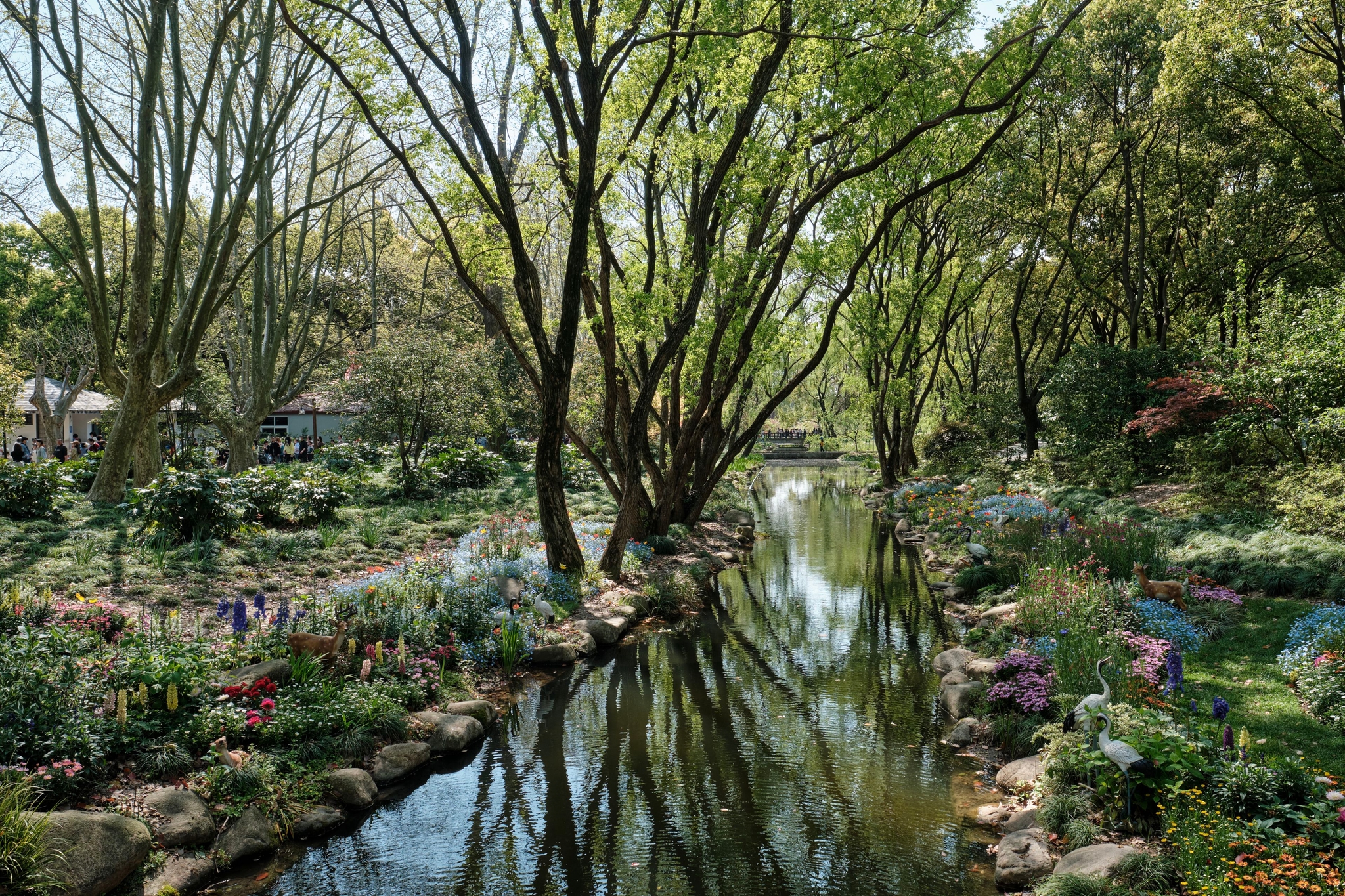 A narrow stream flows through a dense forest or garden with various trees, green foliage, and colorful flowers on both banks. The water reflects the surrounding vegetation
