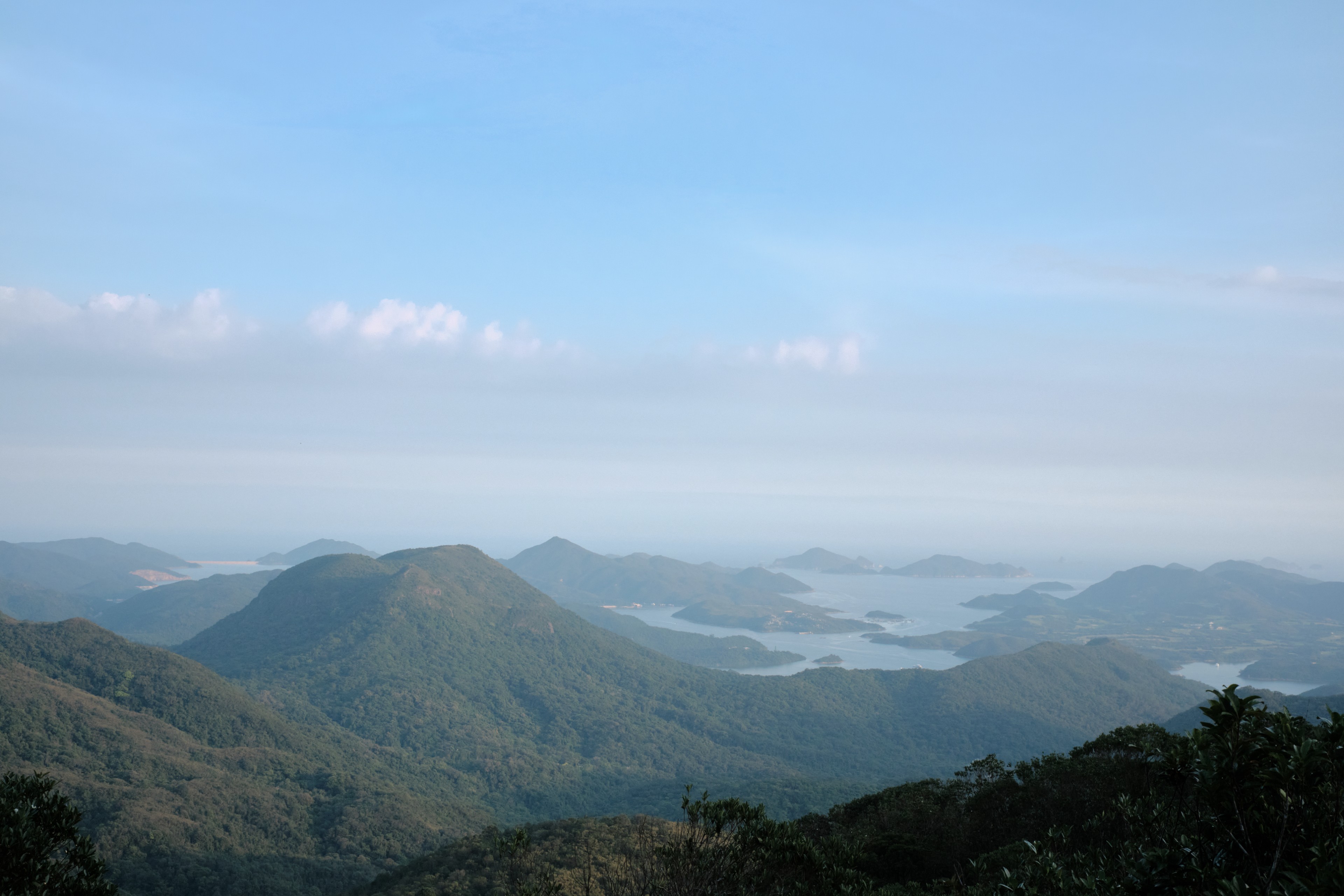 Panoramic view of layered green mountain ranges, hazy distant lake with islands, clear blue sky