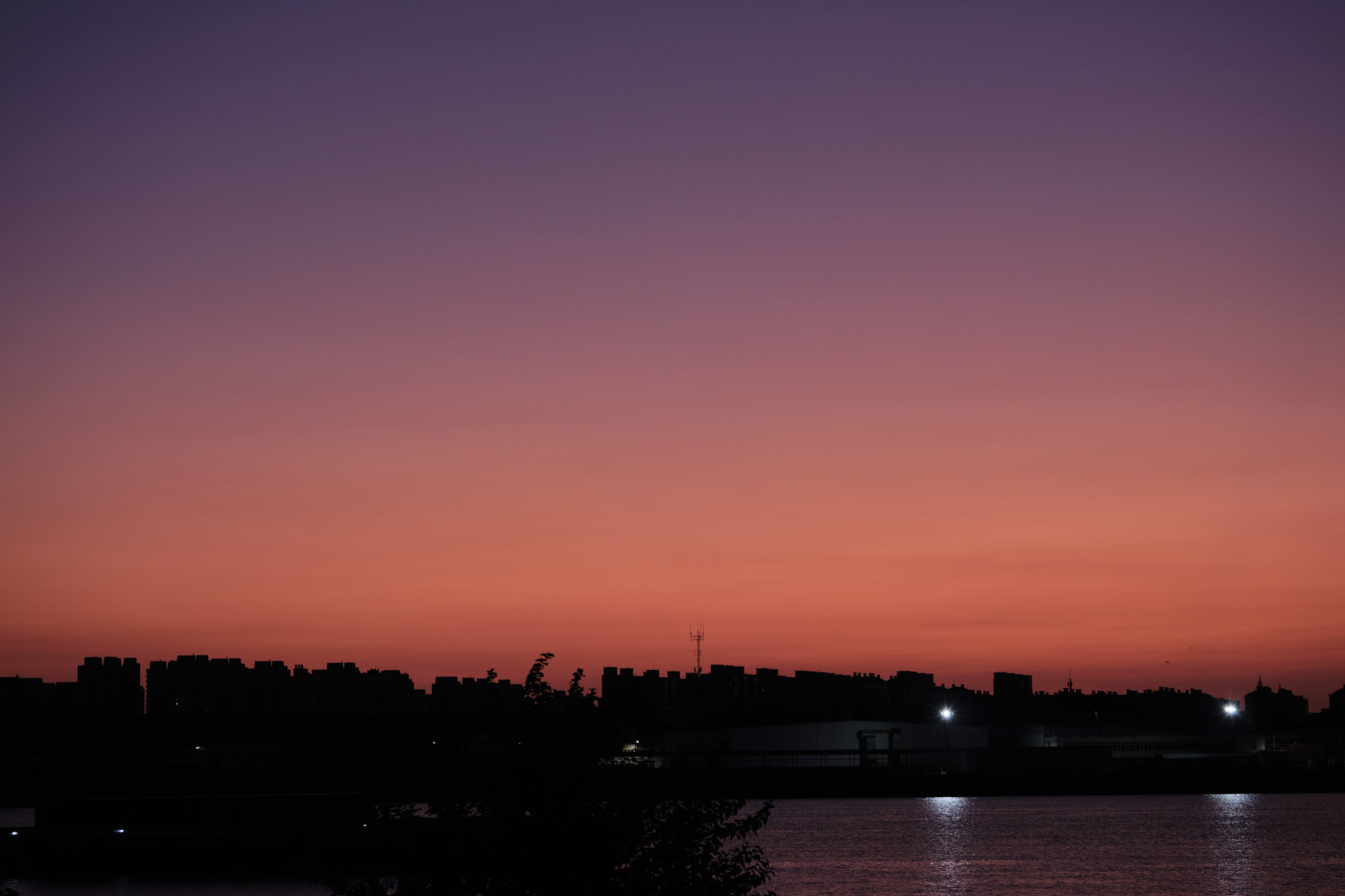 Sky transitions from dark purple to orange-red, silhouetted treeline and distant lights along the horizon, water in the foreground reflects light
