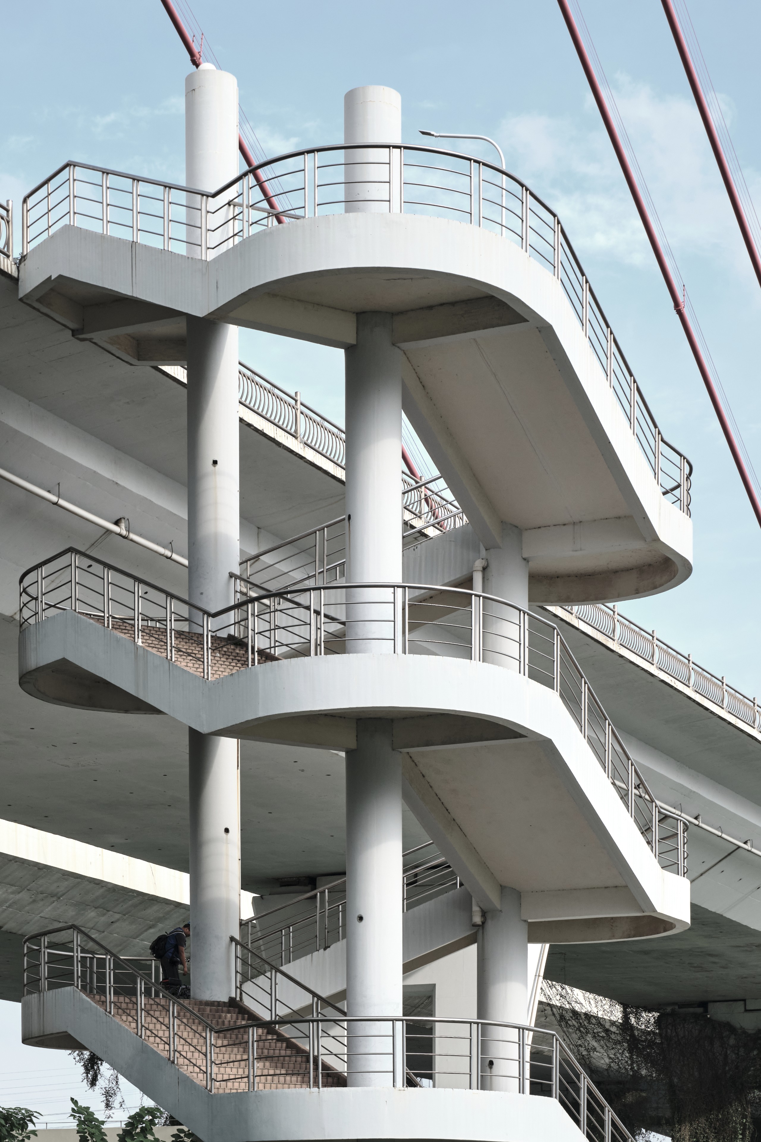 Multi-level concrete spiral pedestrian walkway beneath a bridge, featuring white pillars, metal railings, and grey stairs. A person is visible on the lowest steps. Red suspension cables are in the background against a light blue sky