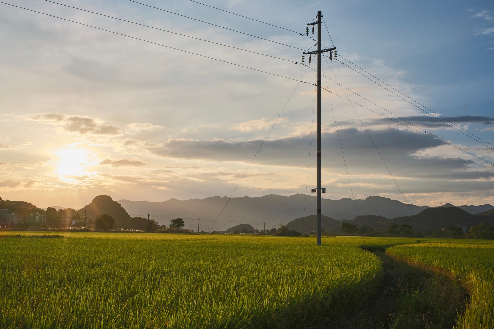 Green field with tall utility pole, setting sun, mountains, and cloudy sky